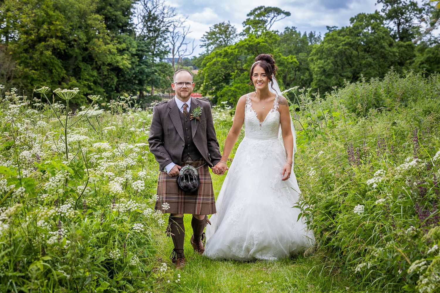 Bride and groom holding hands walking through lush green meadow, groom wearing traditional Scottish kilt.