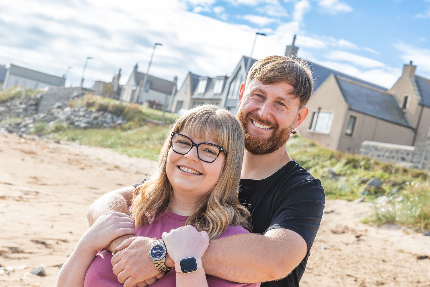 Happy couple smiling outdoors on a sandy area with houses in background on a sunny day.