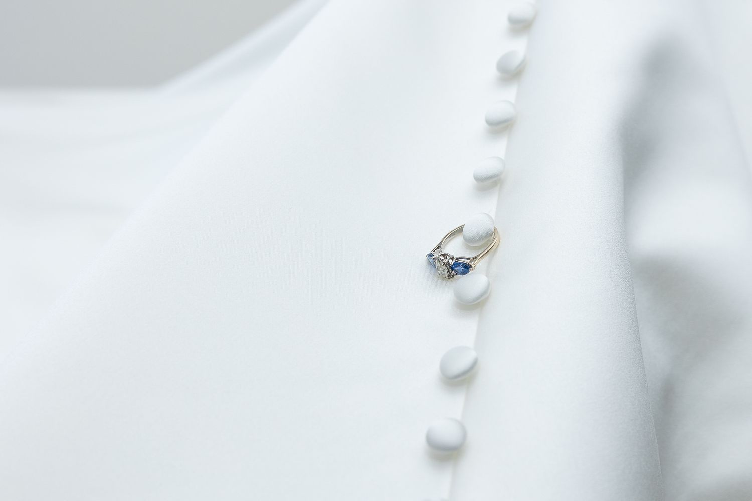 Gold ring with blue sapphire and diamond accents pinned to a white fabric garment with button details.