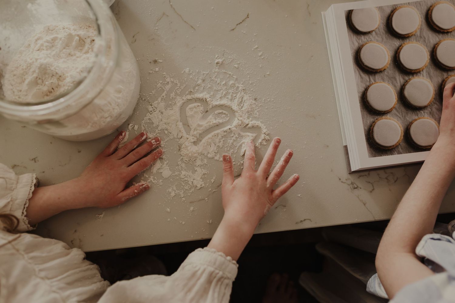 Children baking with flour on a table, a heart drawn in flour beside a muffin tin with cookies.