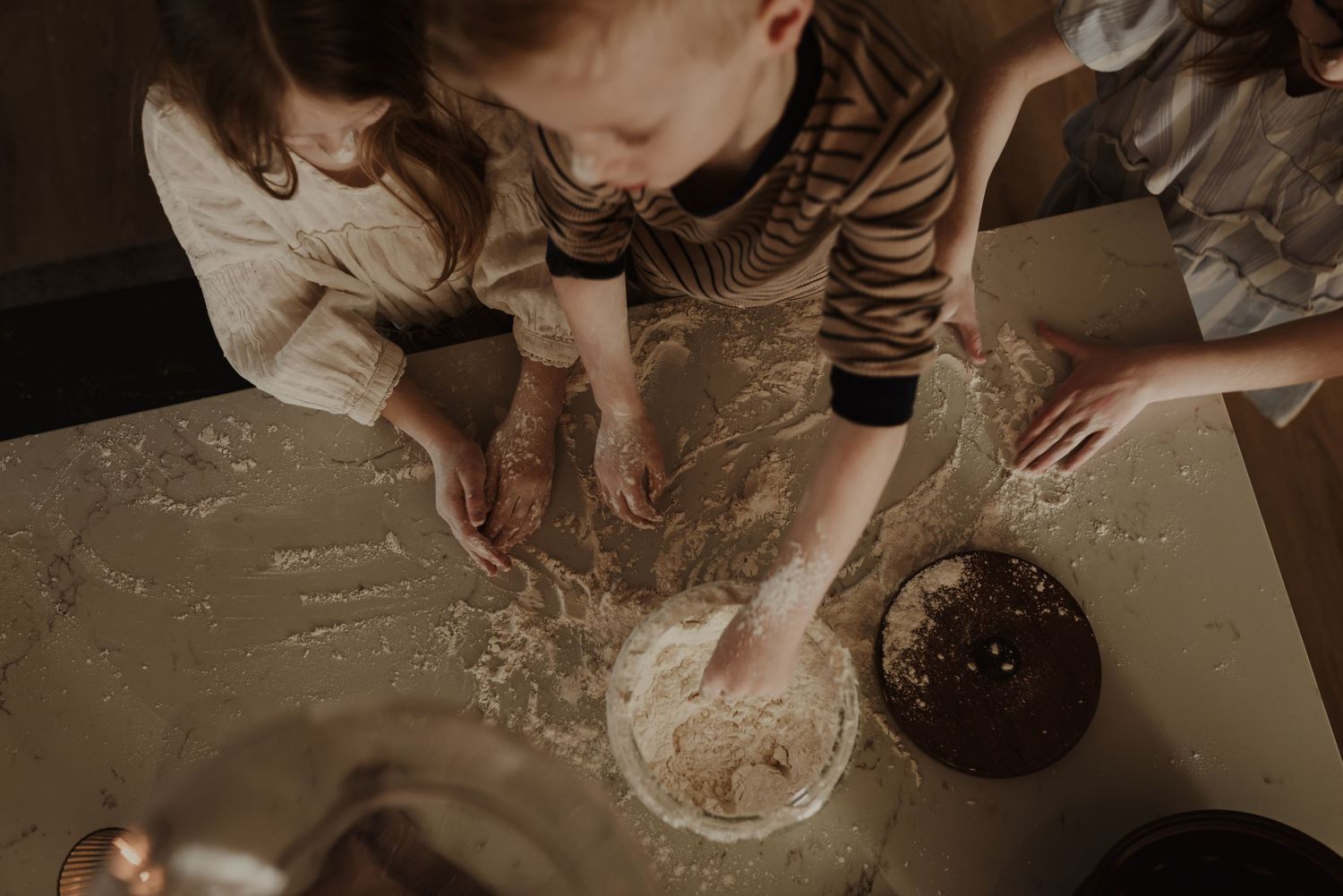 Children knead dough together on a flour-dusted table, learning to bake in a warm, rustic kitchen setting.