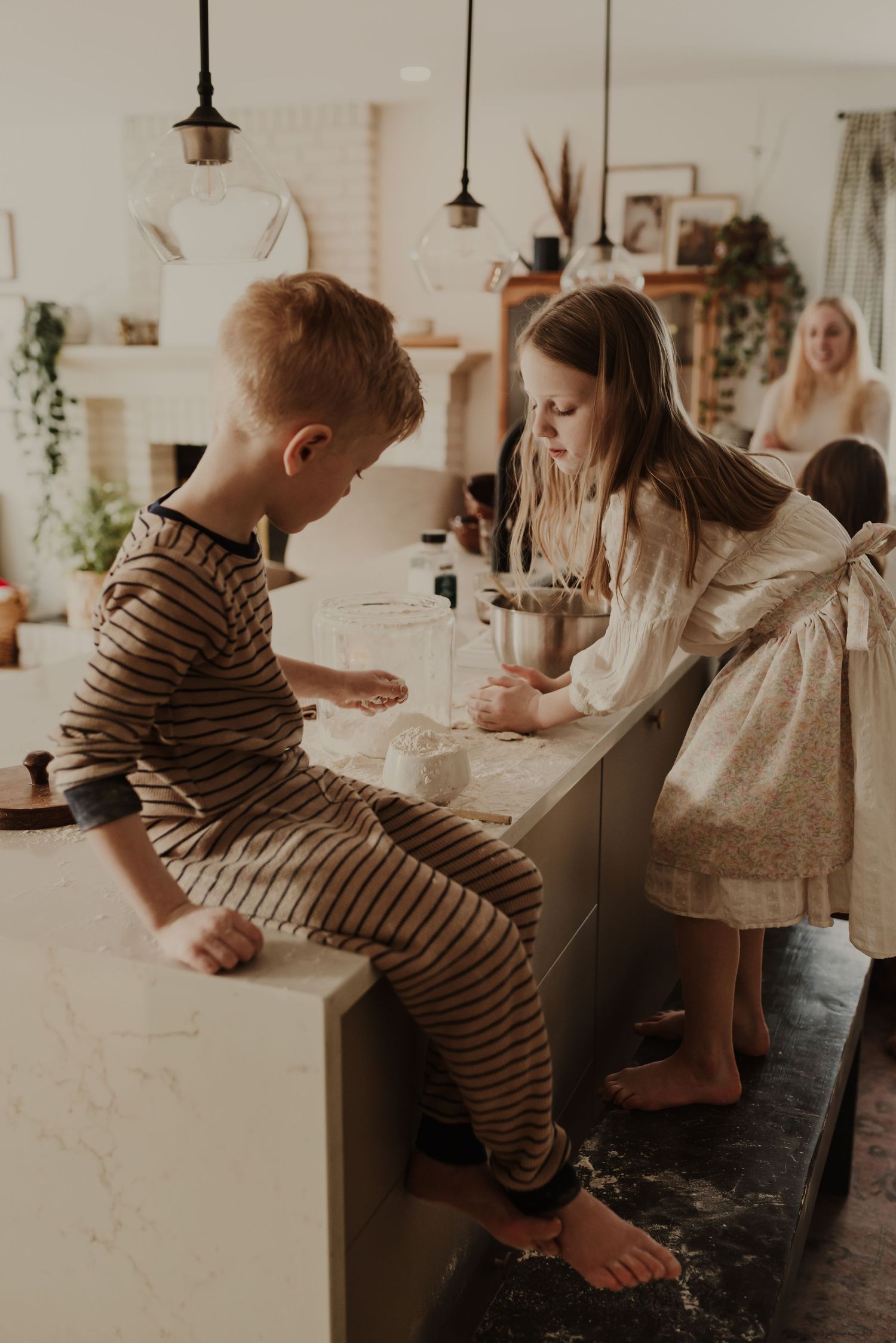 Two barefoot children baking together at a kitchen island, with a woman visible in the cozy background.