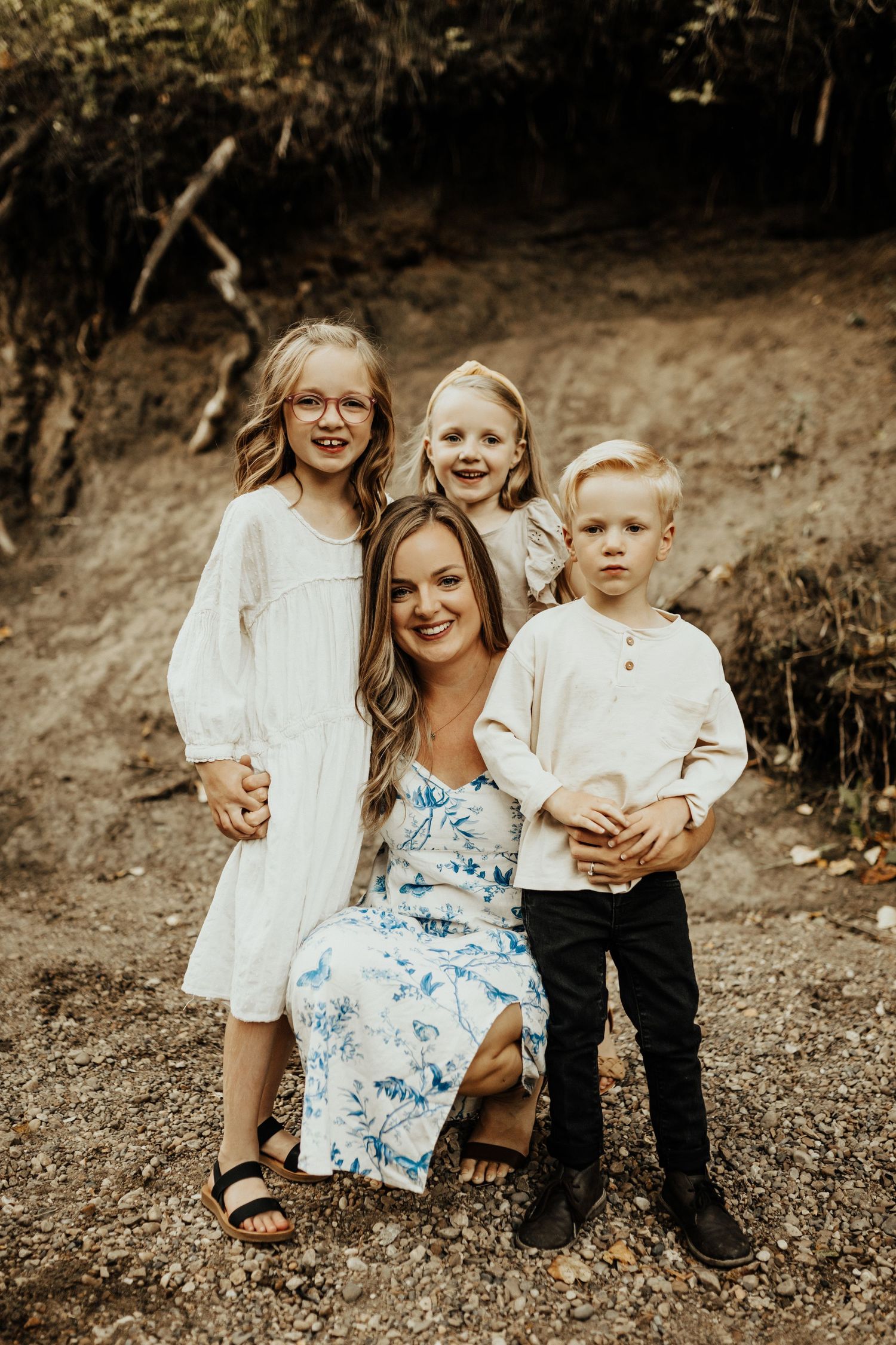 A smiling woman in a floral dress poses outdoors with three young children in white and neutral outfits.
