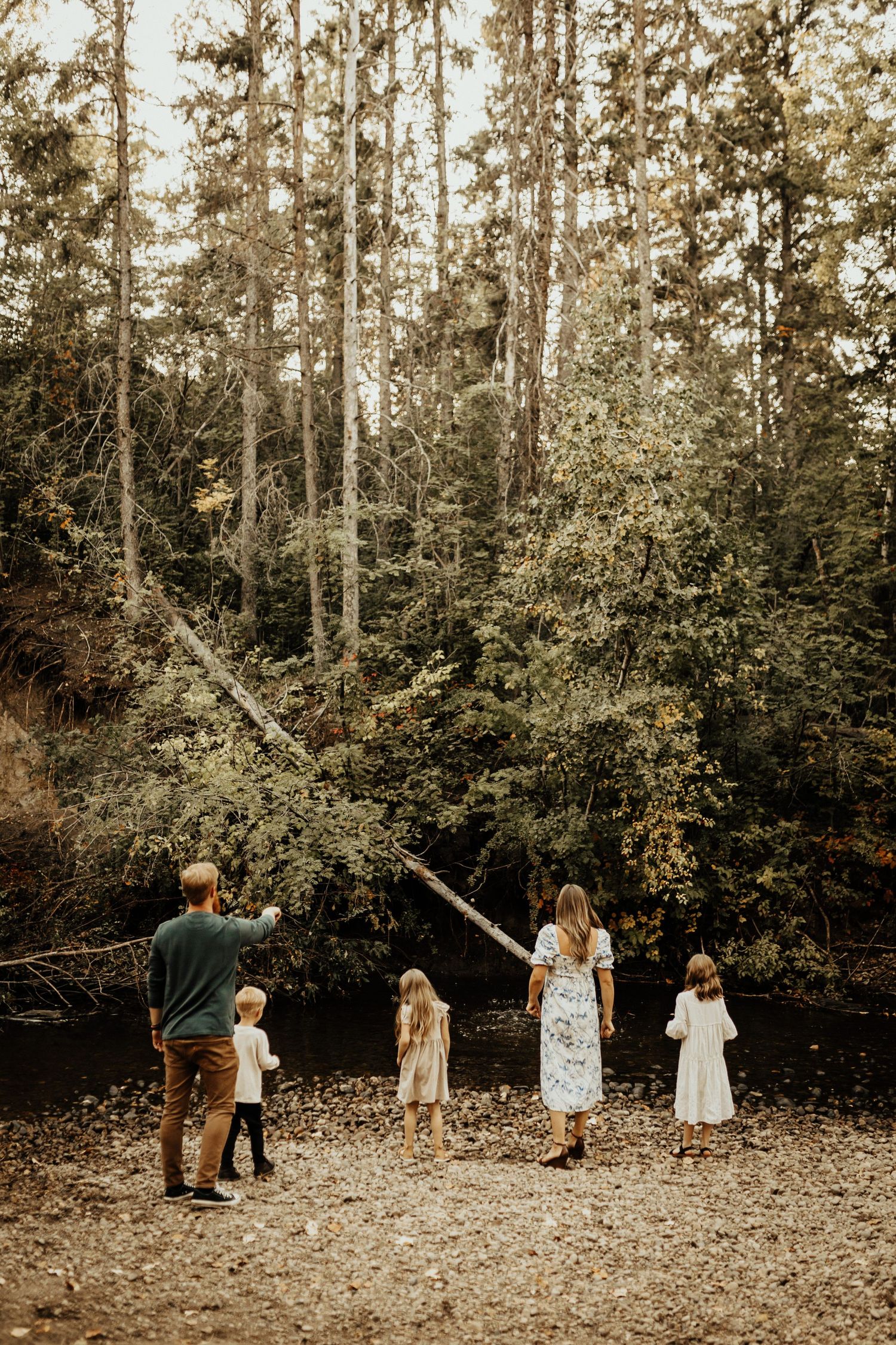 Family of five standing on rocky shore facing lush forest, parents pointing toward trees with children in white dresses.