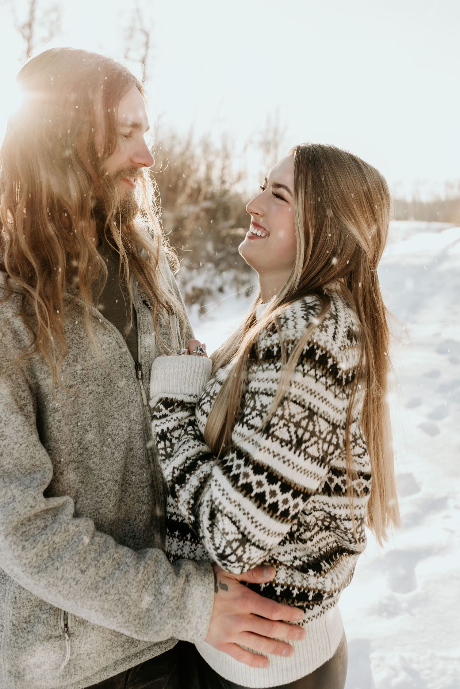 Couple embracing and laughing together in a snowy winter landscape, wearing cozy sweaters with sunlight glowing behind them.