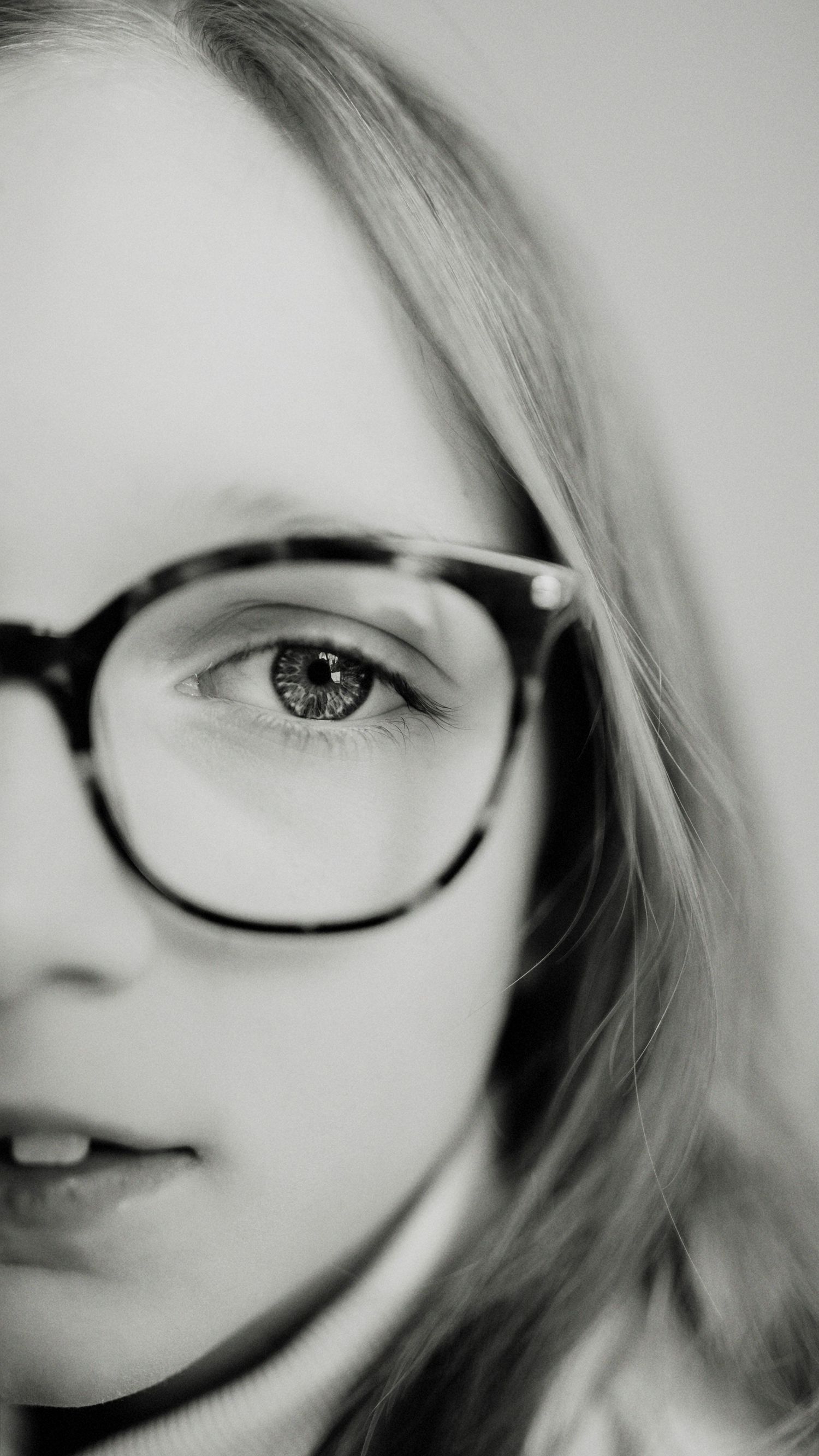 Black and white close-up portrait of a young blonde girl wearing oversized round tortoiseshell glasses.