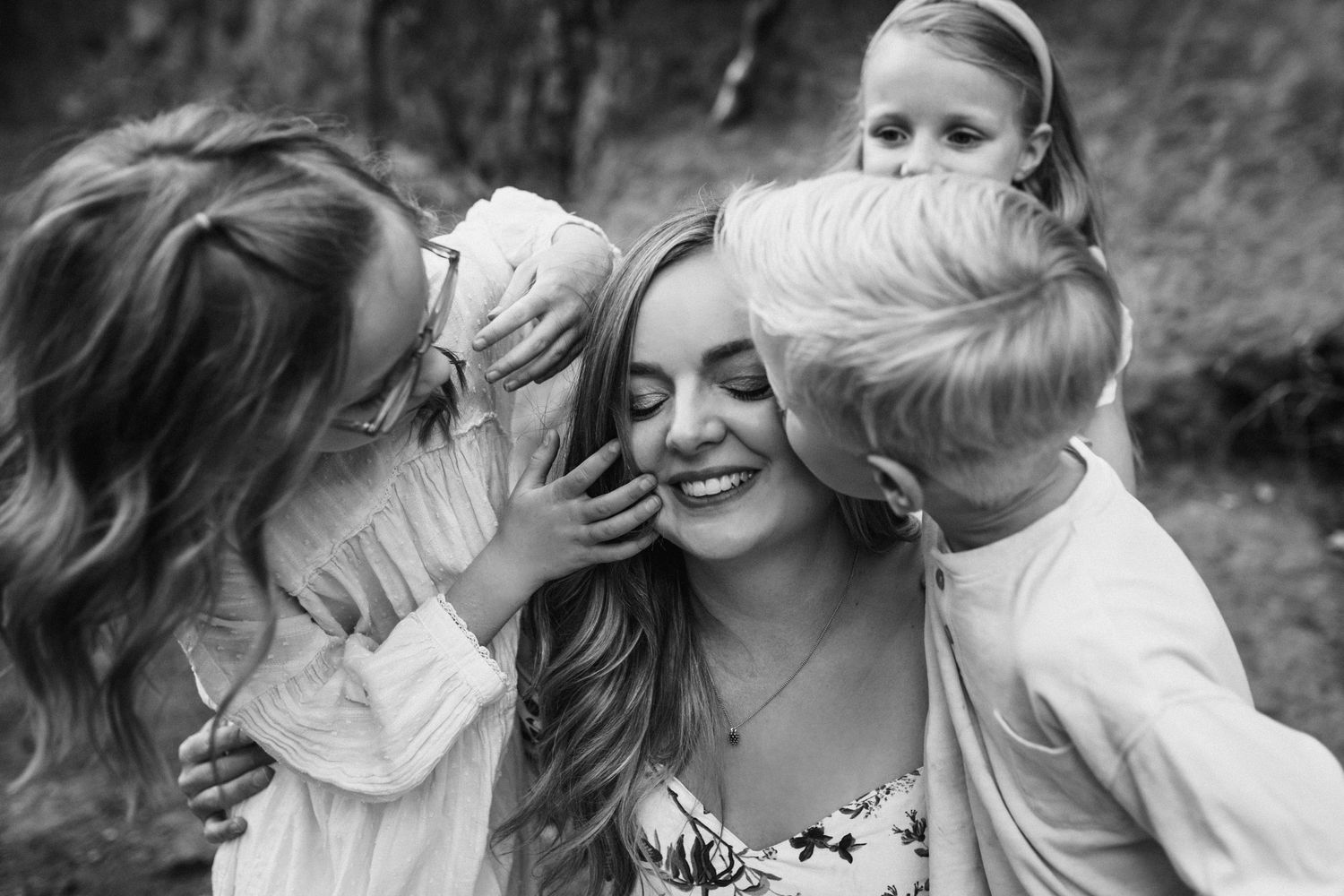 Black and white photo of a smiling woman being kissed on cheeks by two young children outdoors.