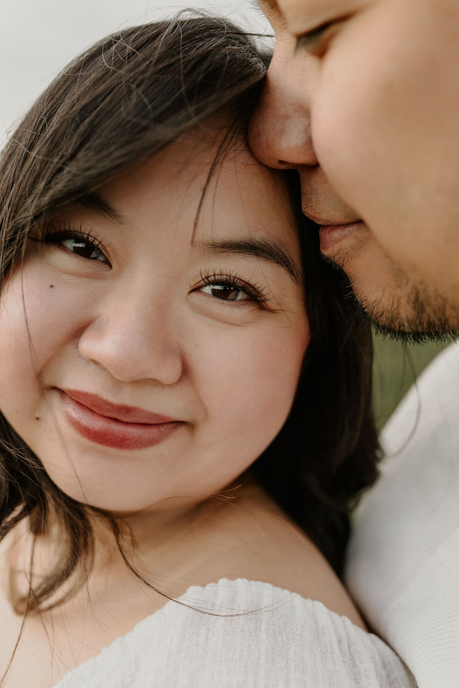 Close-up of a smiling Asian woman as her partner gently kisses her forehead in a romantic couples portrait.