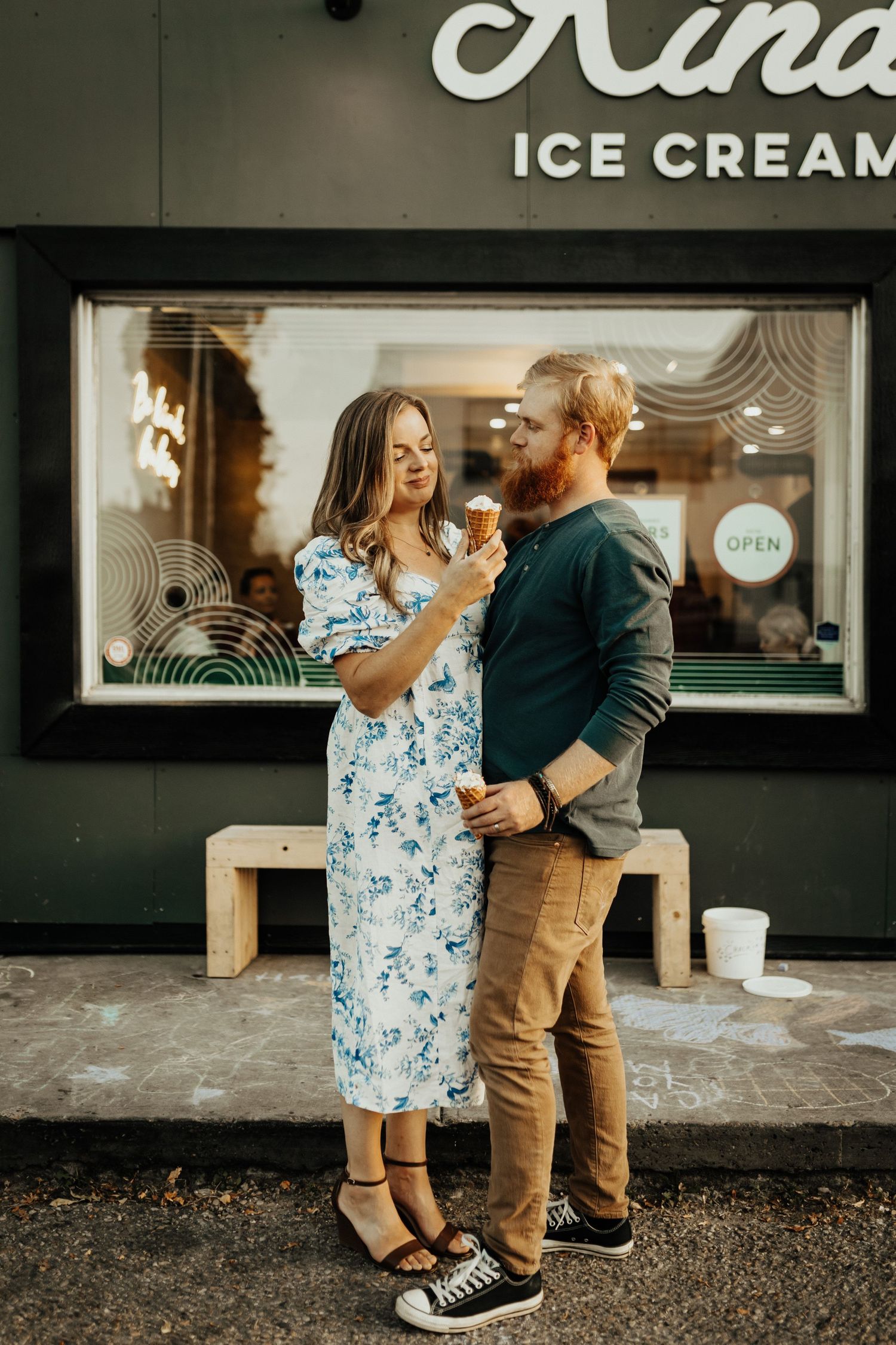 Couple sharing ice cream cones outside a shop, woman in floral dress, man in green sweater and khakis.