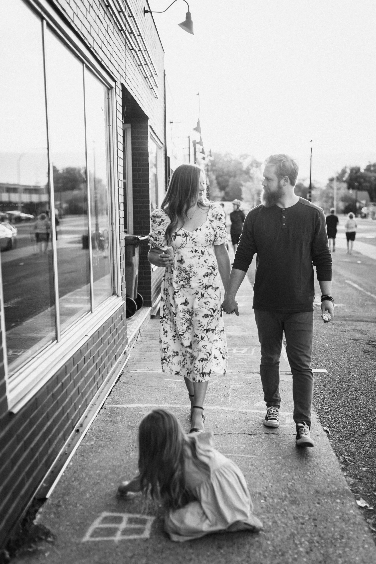 Black and white photo of a couple holding hands on a sidewalk while their child plays behind them.