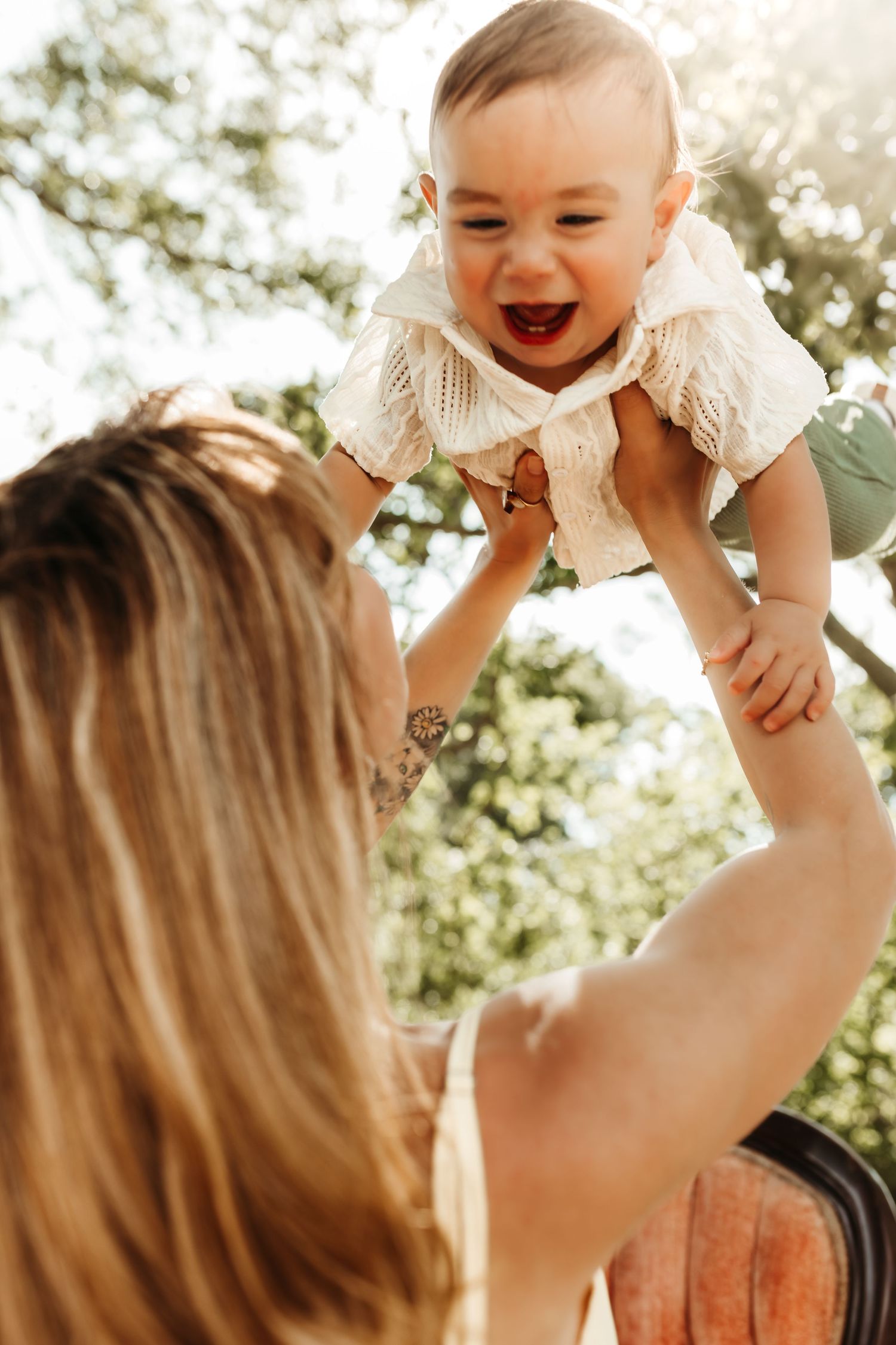 A joyful parent lifts and plays with their laughing baby outdoors in warm sunlight.
