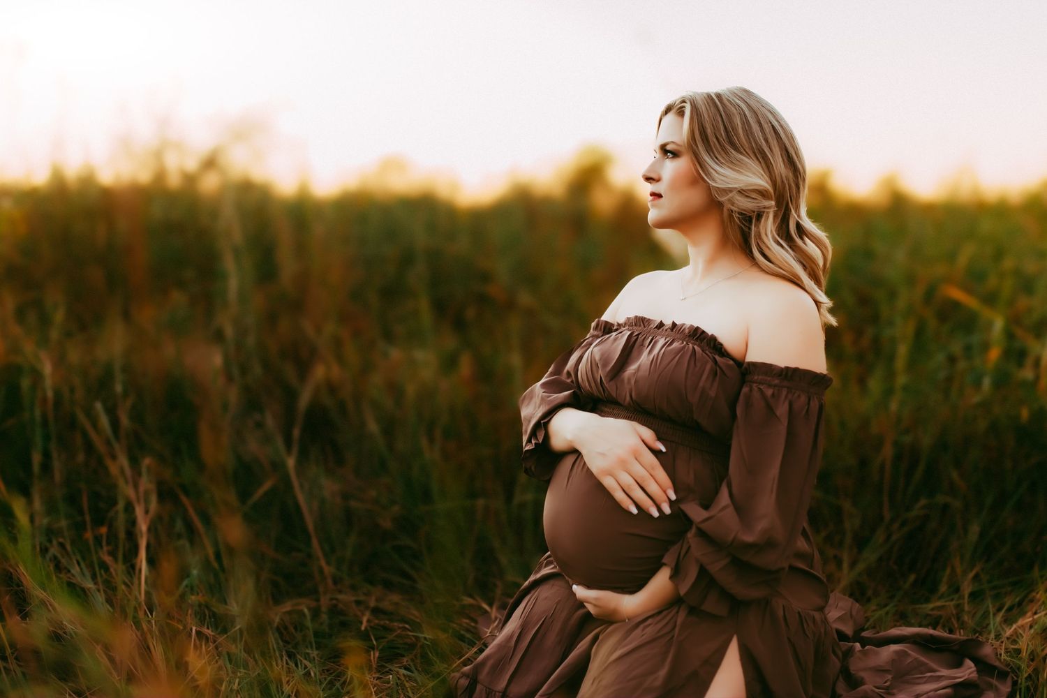 Expecting mother wearing brown off-shoulder dress poses in golden field at sunset.
