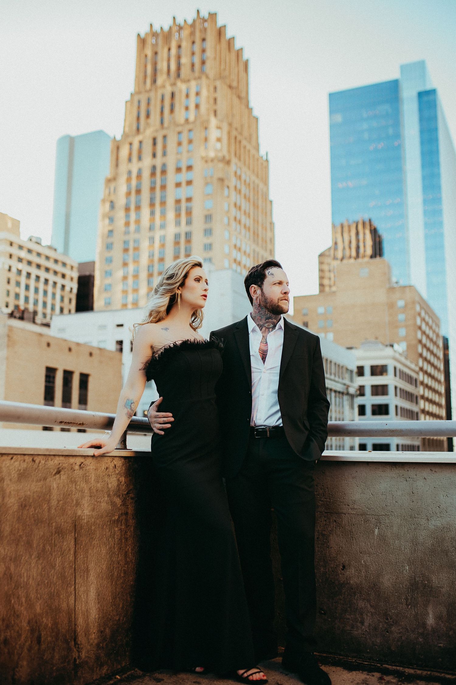 Stylish couple in black attire stands on urban rooftop against backdrop of art deco buildings and modern skyscrapers.