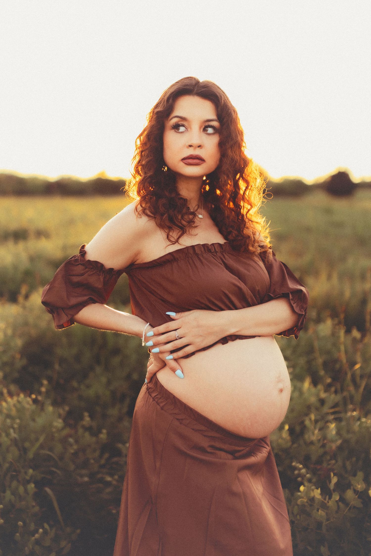 A woman in a brown dress poses in a sunlit meadow during golden hour for a maternity photoshoot.