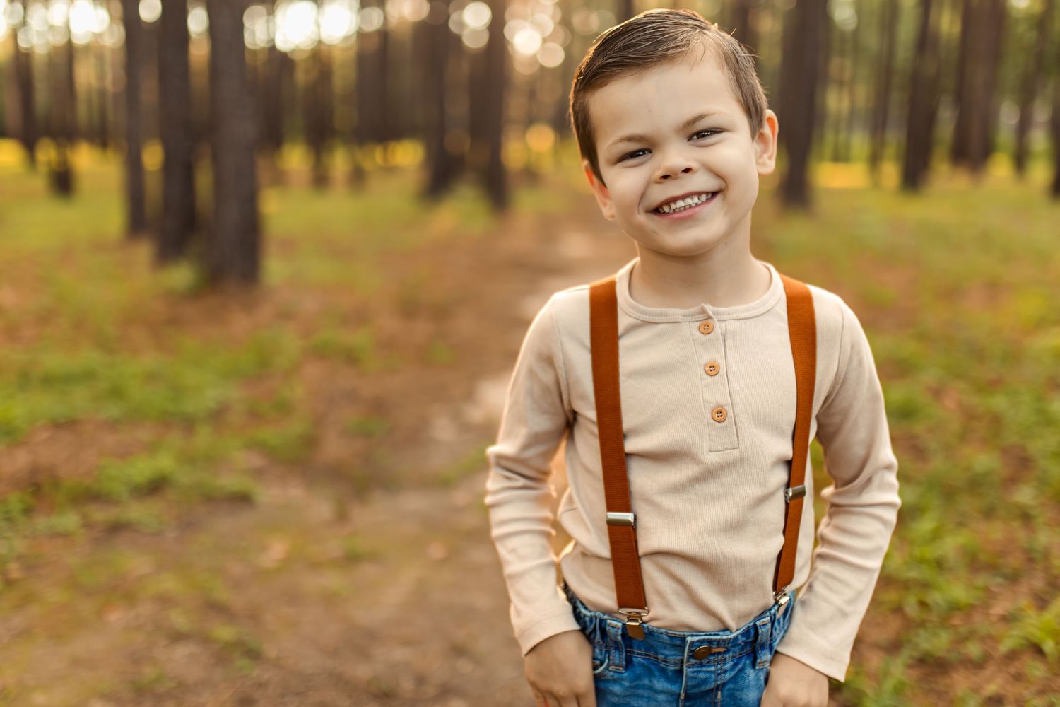 A child in beige shirt and denim overalls with suspenders plays in an autumn forest setting.
