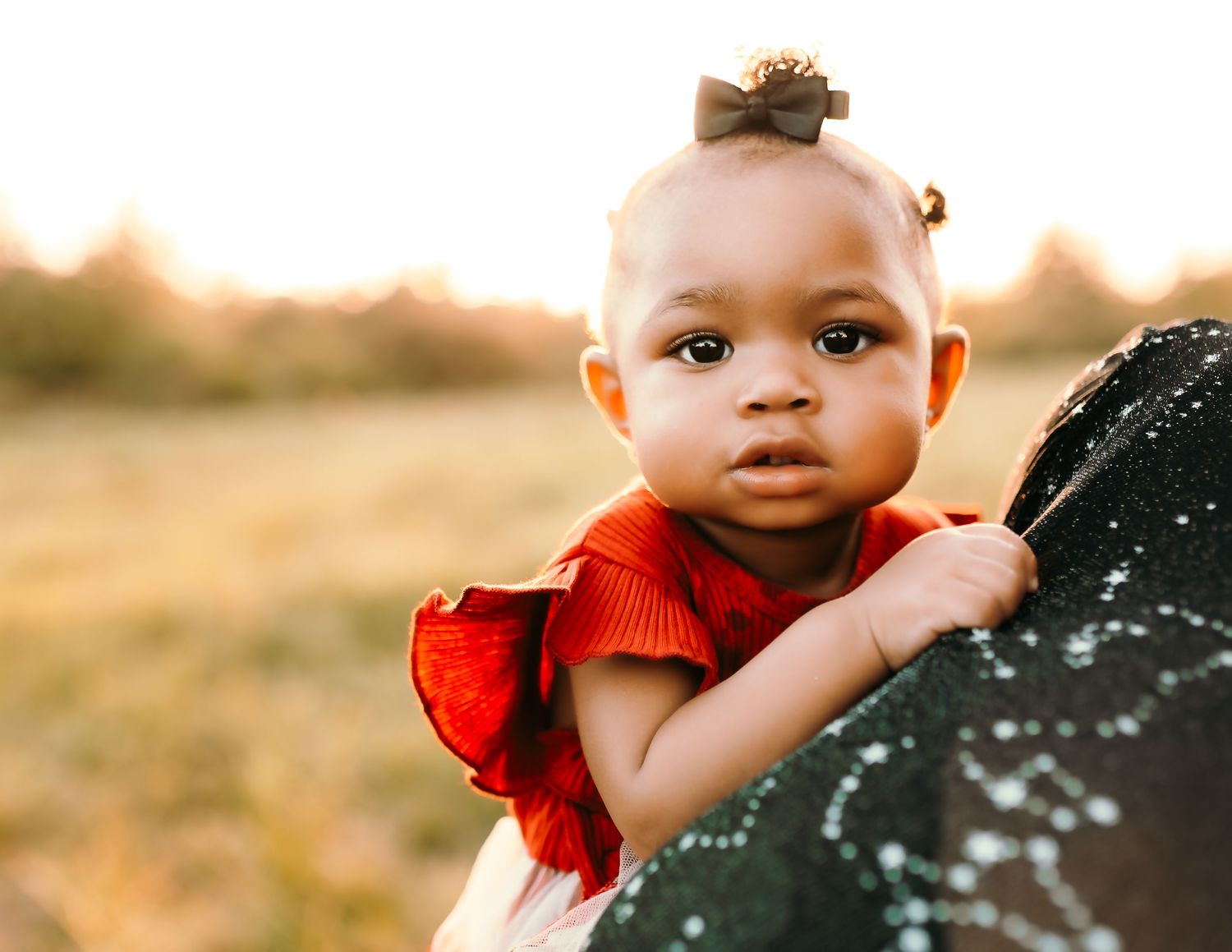 A baby in a red sweater smiles while being held outdoors during golden hour sunset.