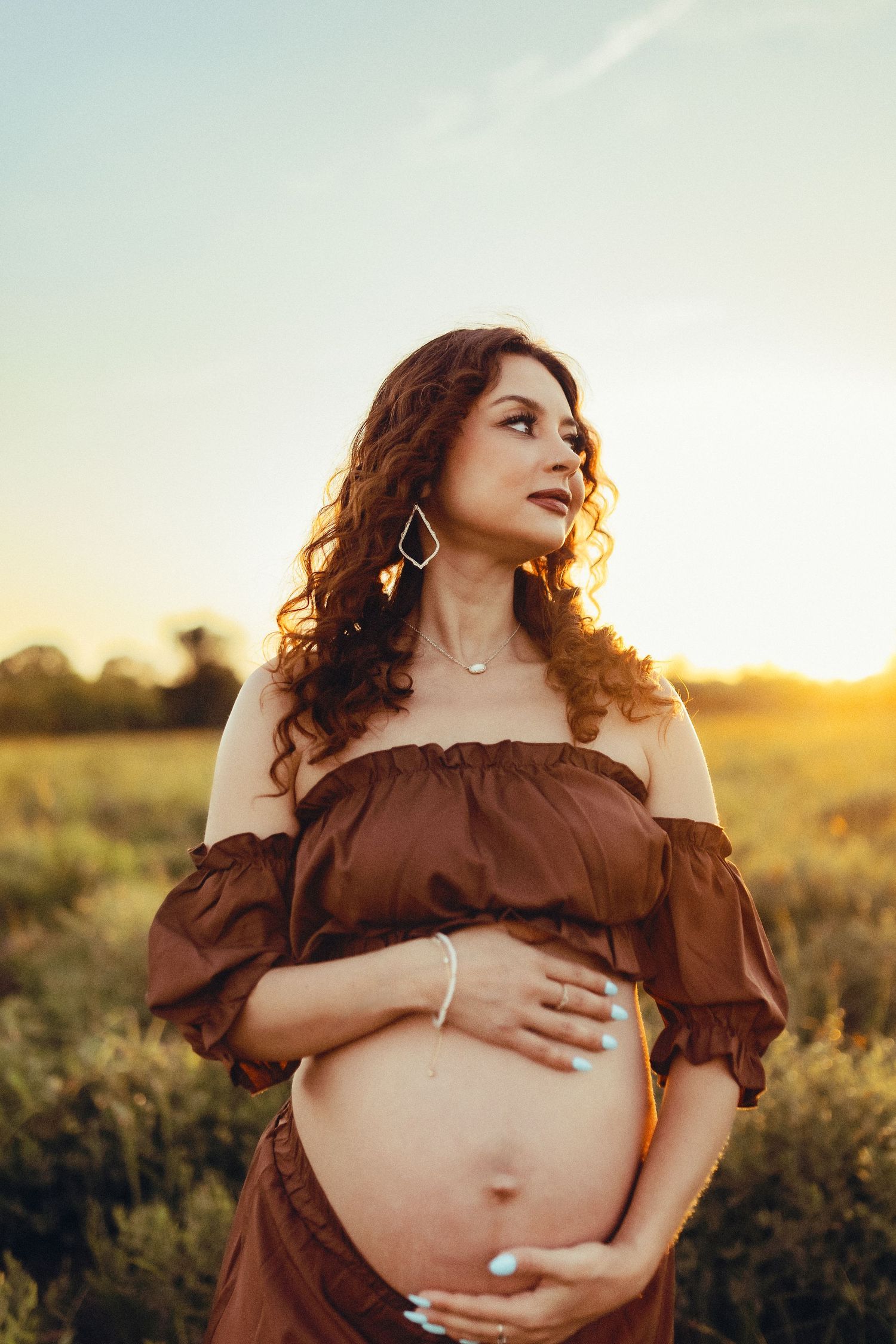 A pregnant woman in a brown off-shoulder dress poses in a golden field at sunset.