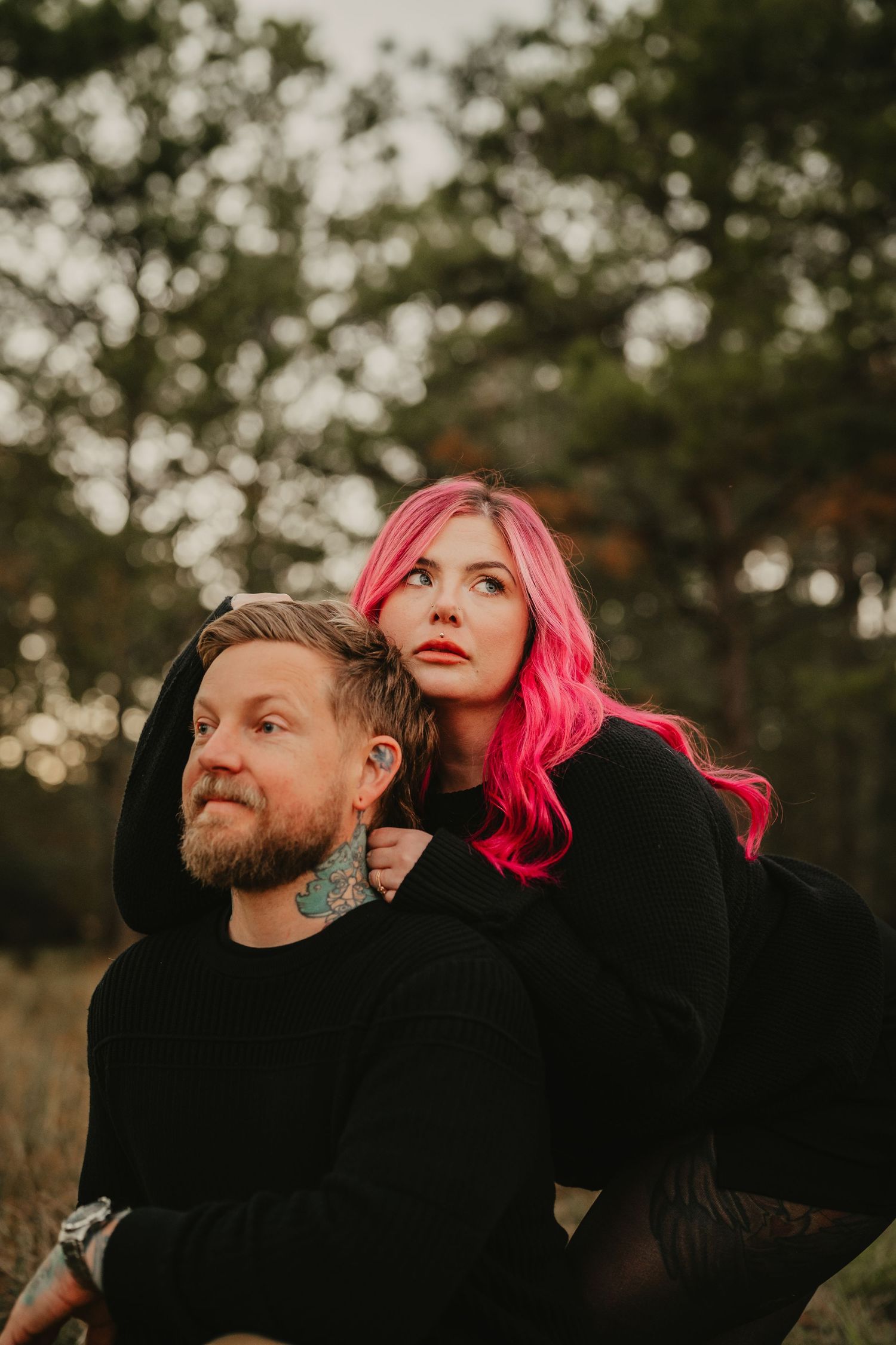 Couple in black outerwear poses together at sunset in moody outdoor portrait with vibrant pink hair detail.
