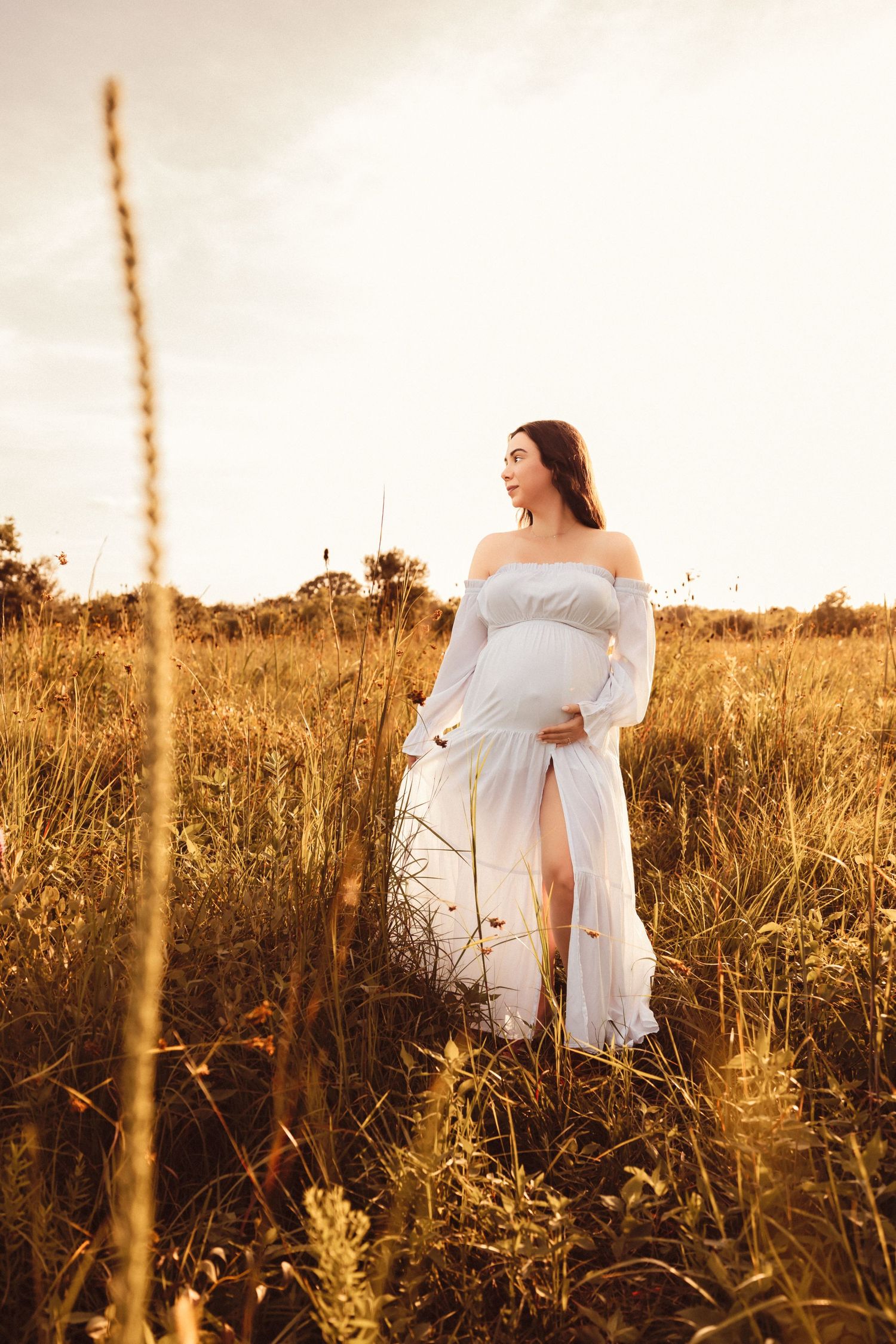 A woman in a flowing white dress stands gracefully in a golden sunlit field during sunset.