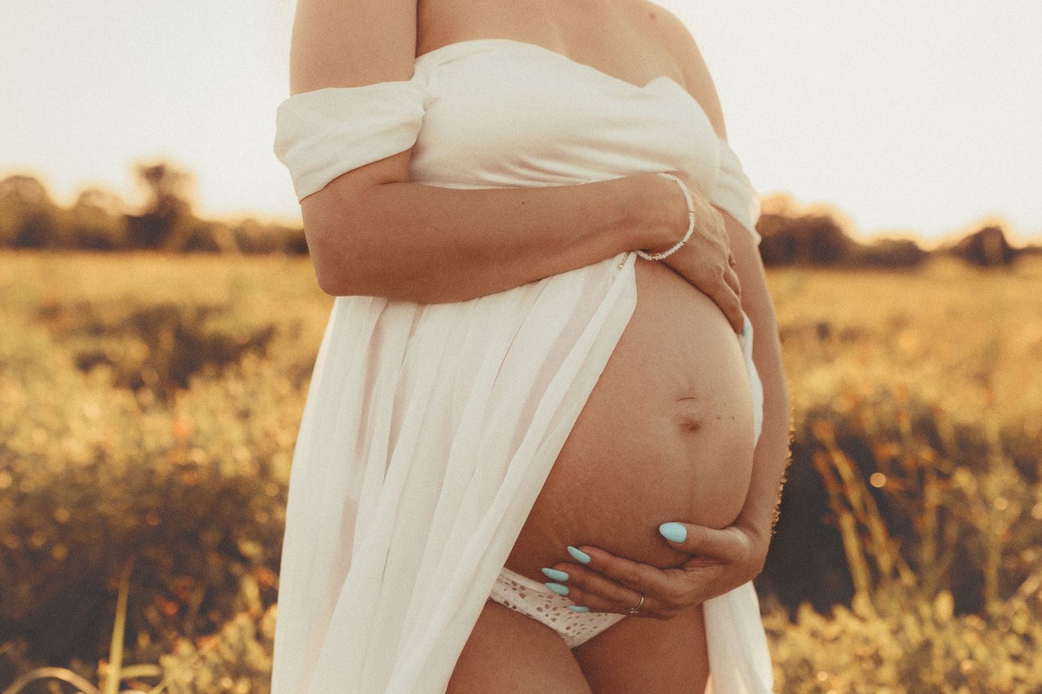 A pregnant belly wrapped in flowing white fabric photographed during golden hour in a sunlit field.