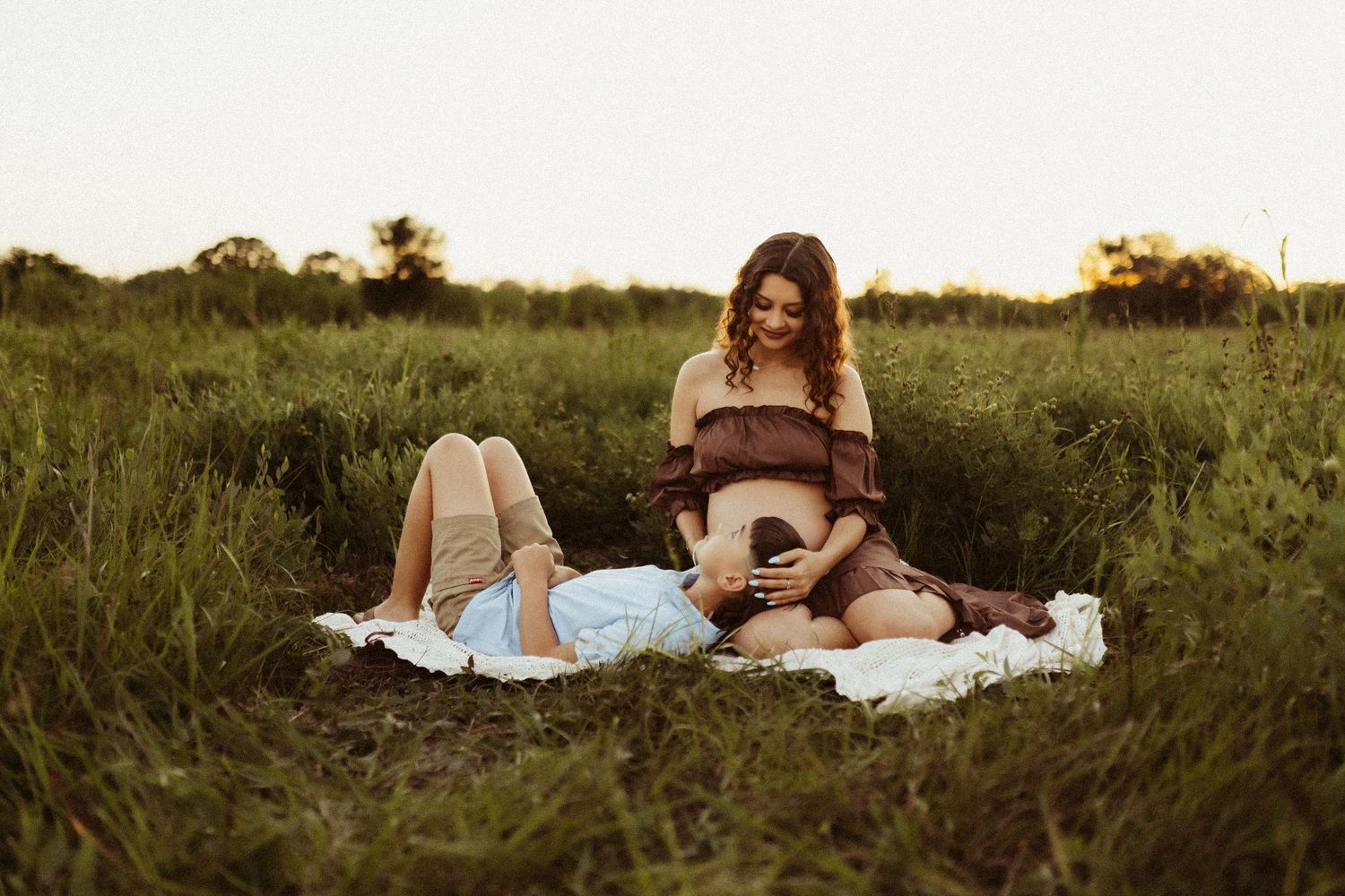 Someone sits on a white blanket in a grassy field at sunset while playing an acoustic guitar and singing.
