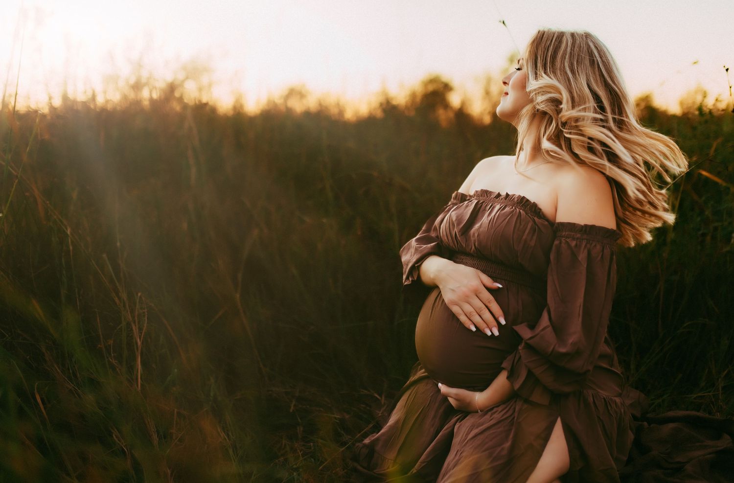 A silhouetted figure in a flowing brown dress tenderly cradles their pregnant belly at sunset in a rustic field.