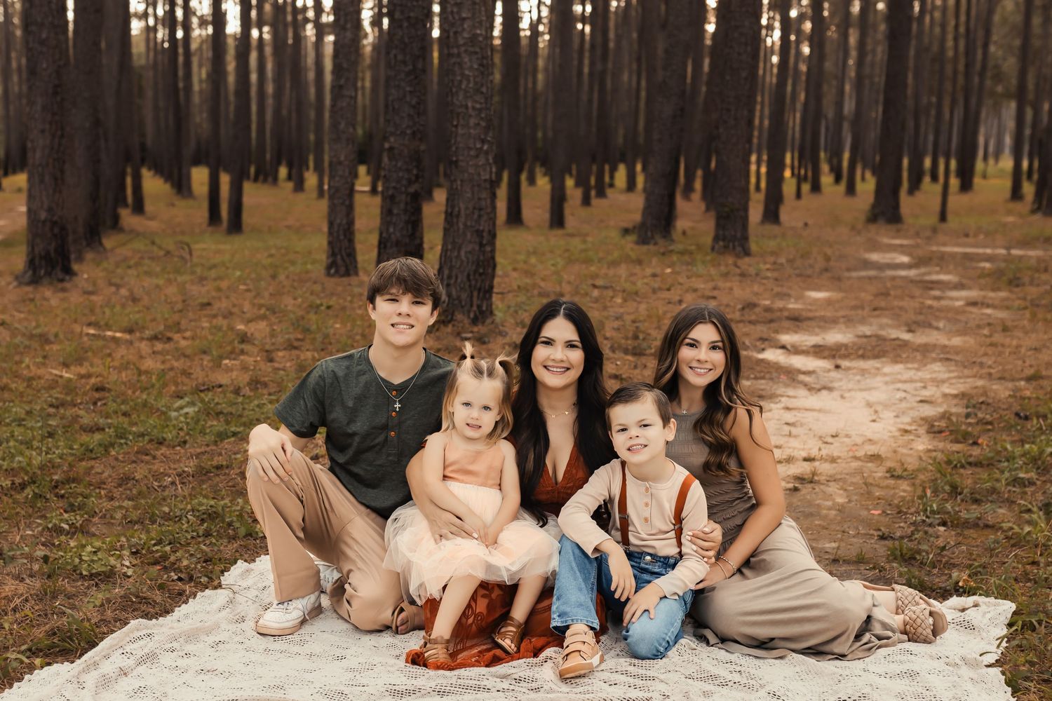 A family sits together on a white blanket in a serene pine forest during golden hour.