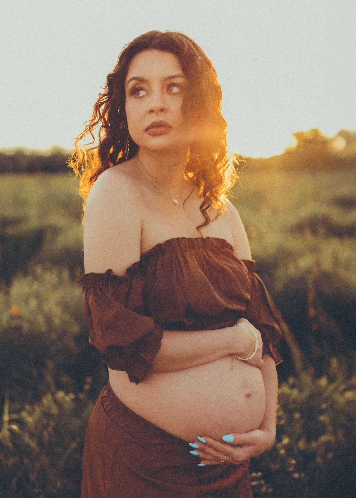 Artistic maternity portrait of figure in brown dress against golden sunset light in natural field setting.