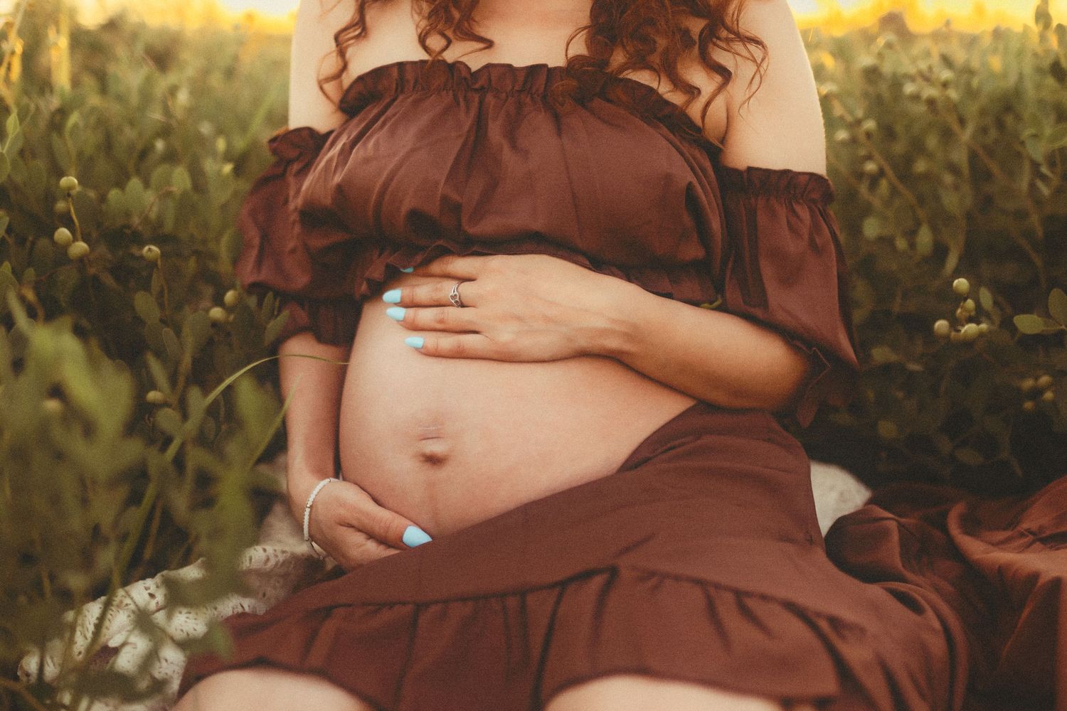 A pregnant figure in a brown dress cradles their belly while sitting in a sunlit garden at golden hour.