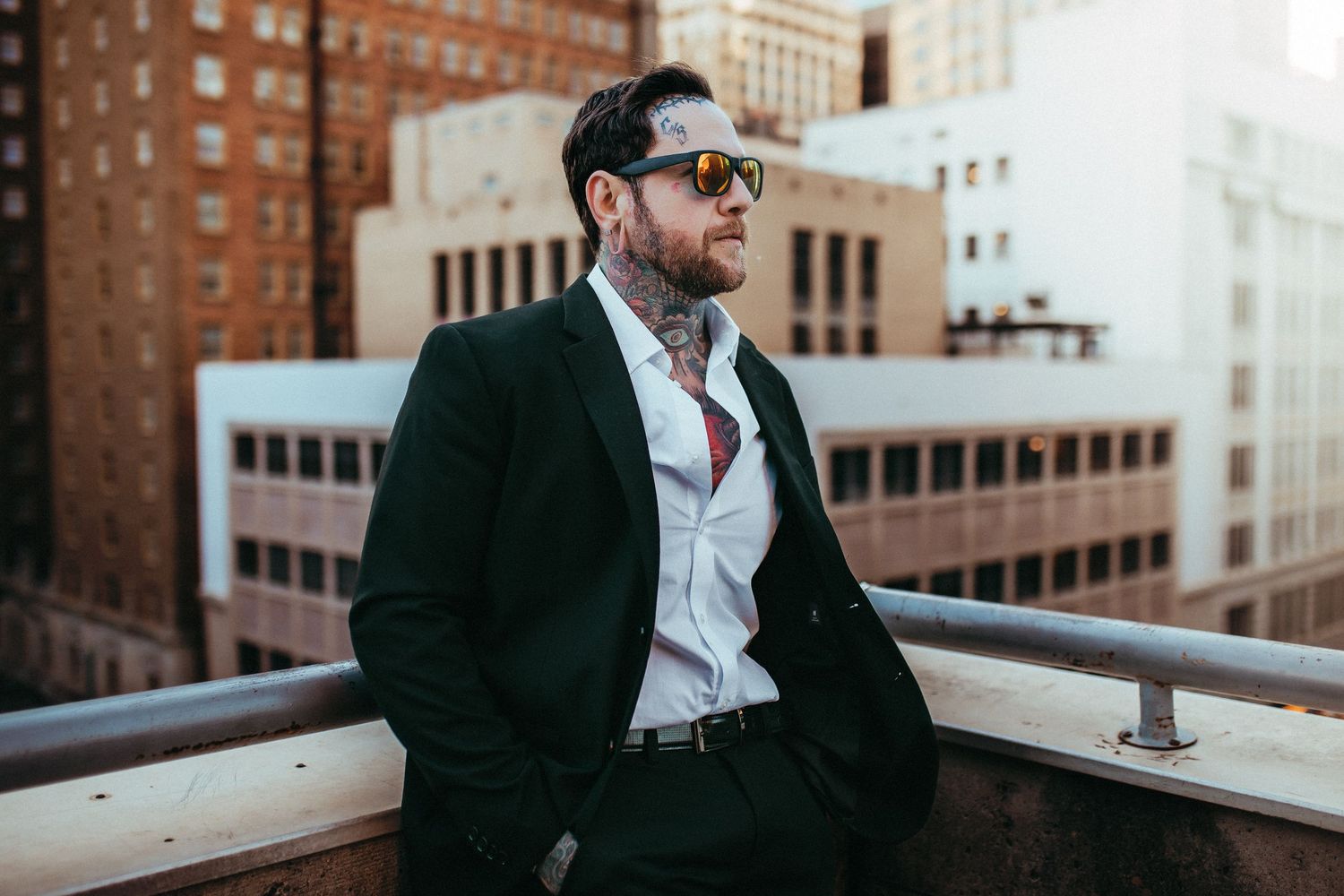 Professional wearing sunglasses and business attire stands on urban rooftop overlooking city buildings.