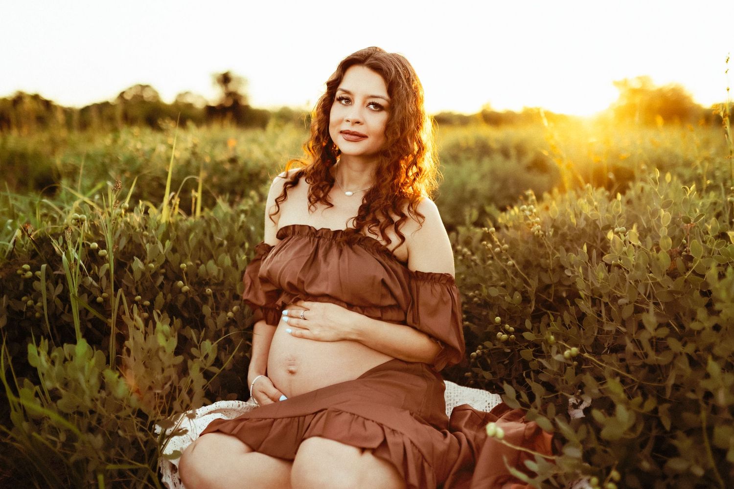 Woman in brown dress poses in golden field at sunset, creating romantic outdoor maternity portraits.