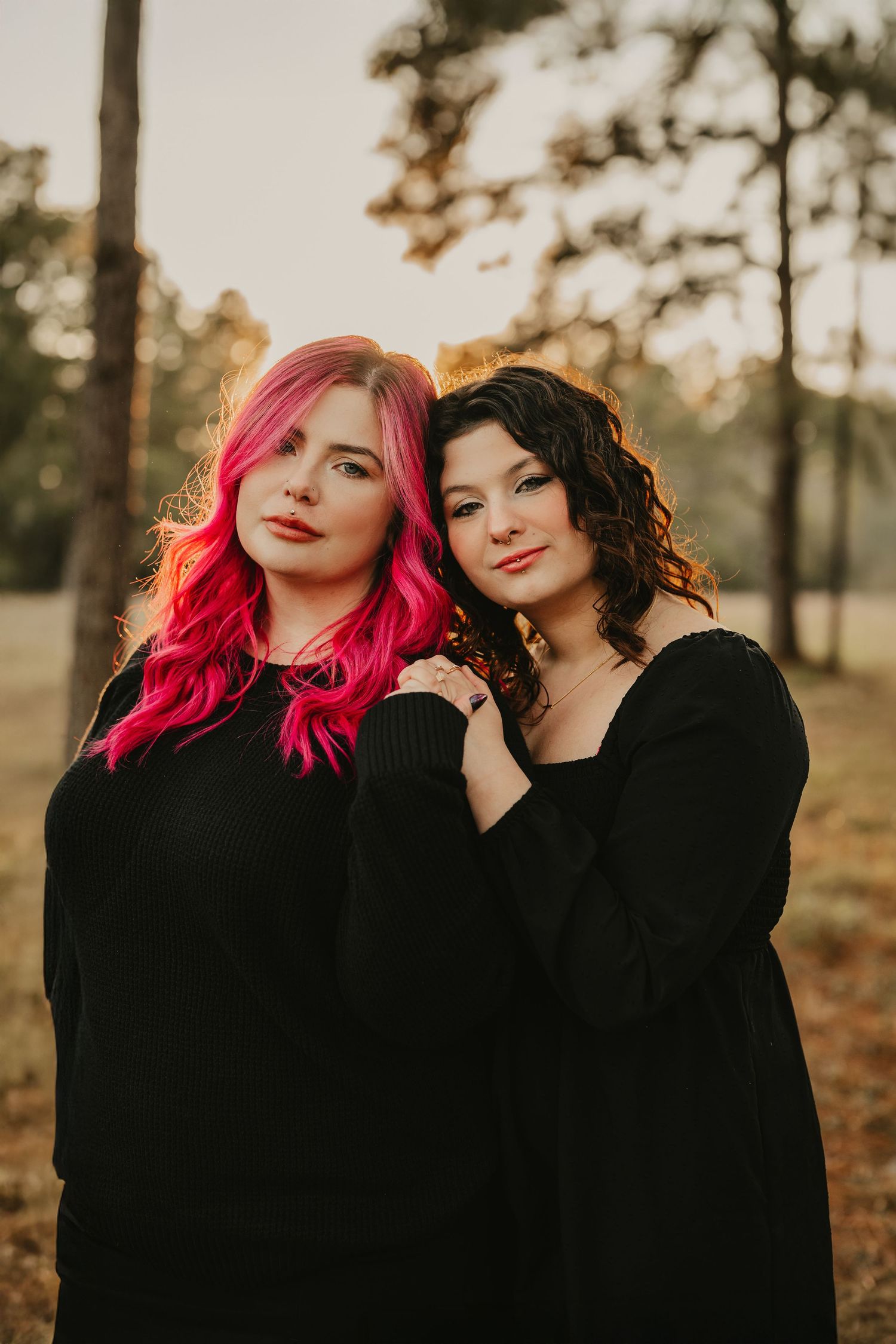 Two women in black dresses pose together during golden hour with one sporting vibrant pink hair.