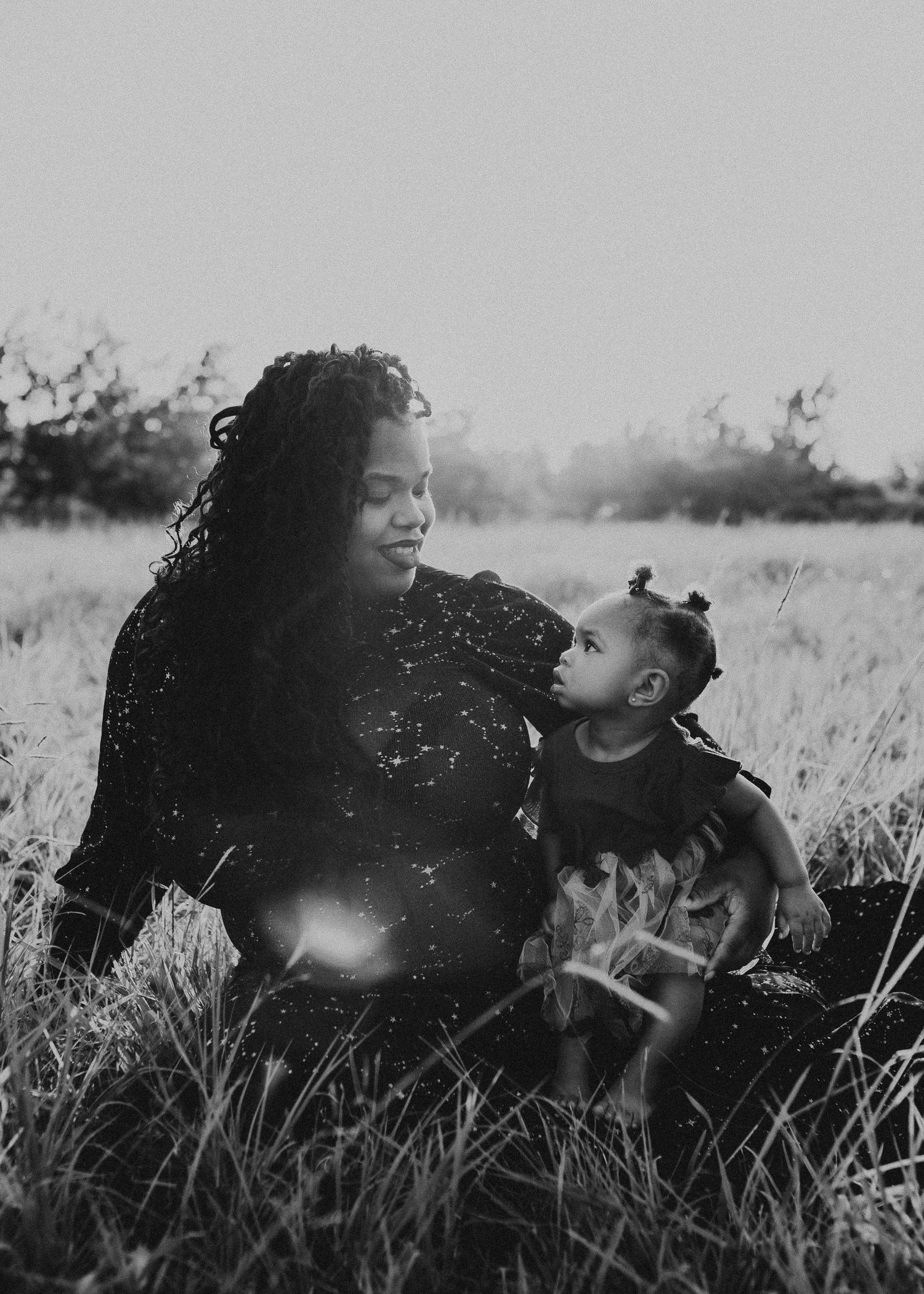 A tender black and white portrait of a parent and child sitting together in a grassy field on a sunny day.
