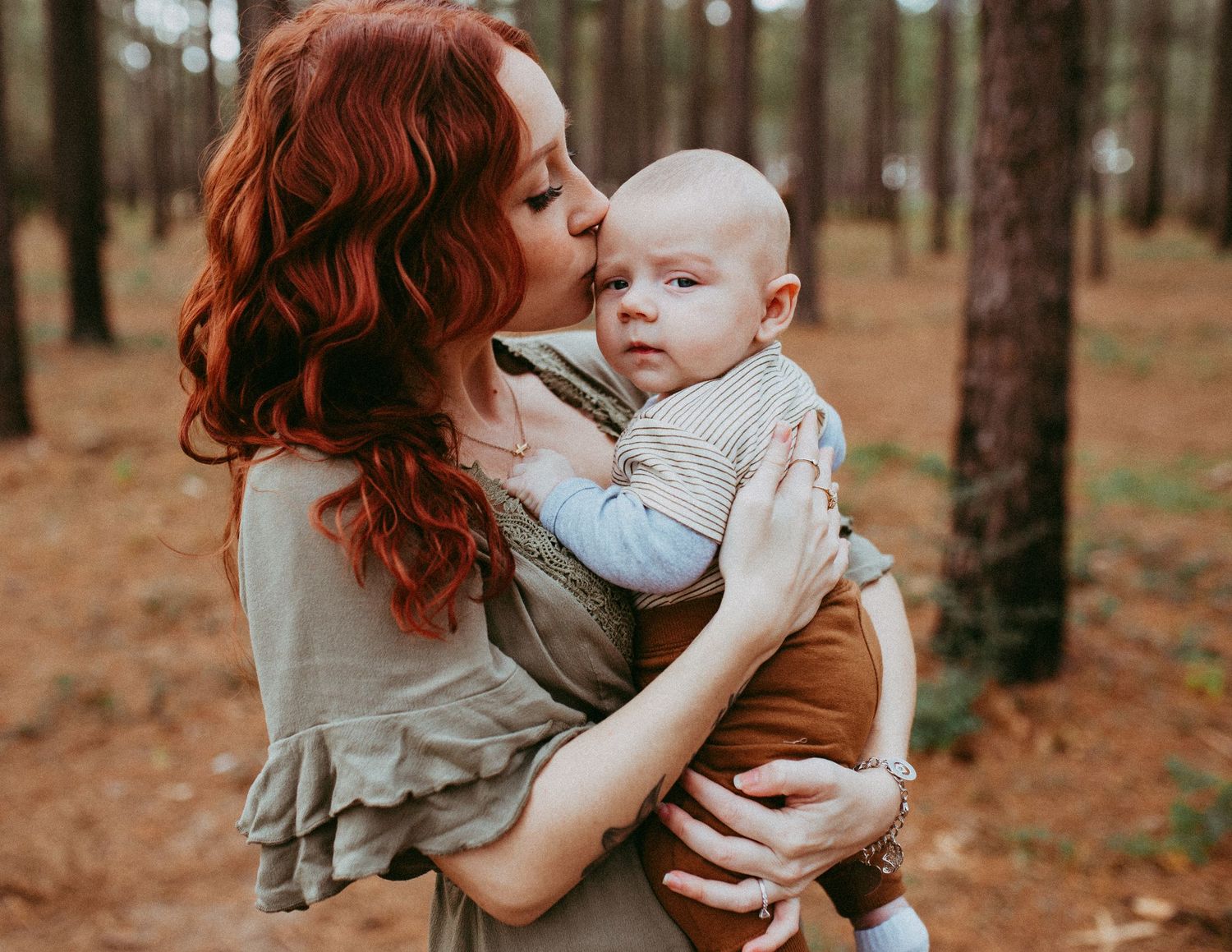 A person in brown clothing holds a baby in a gray onesie while standing among tall pine trees in a forest setting.
