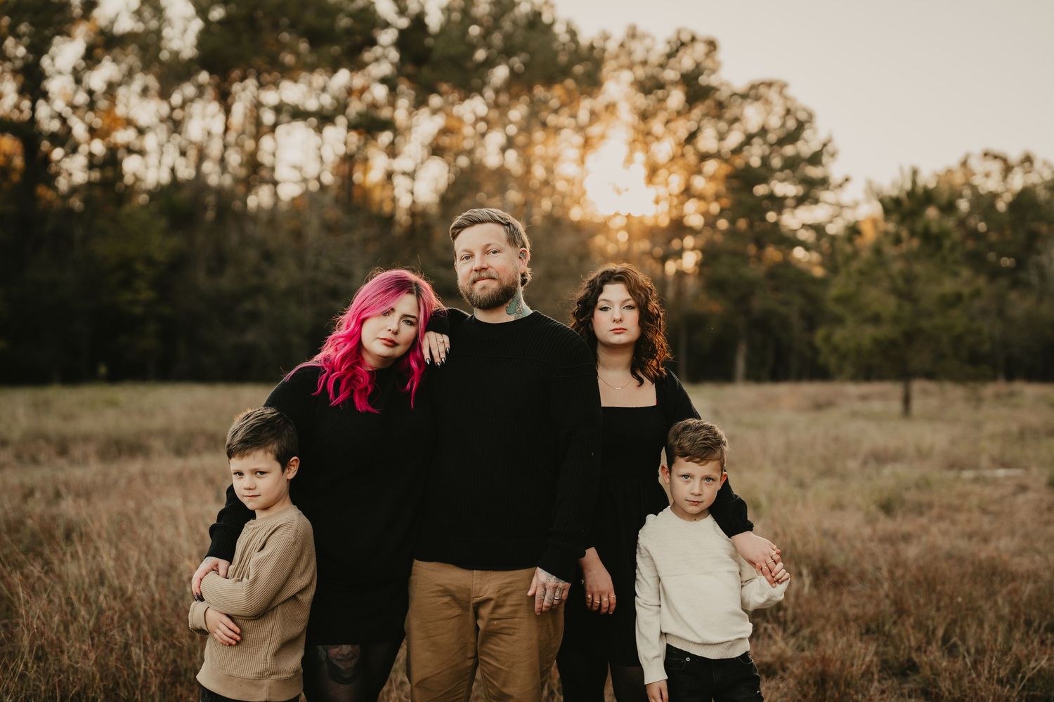 Candid family portrait of five people standing together in a rustic field at sunset.