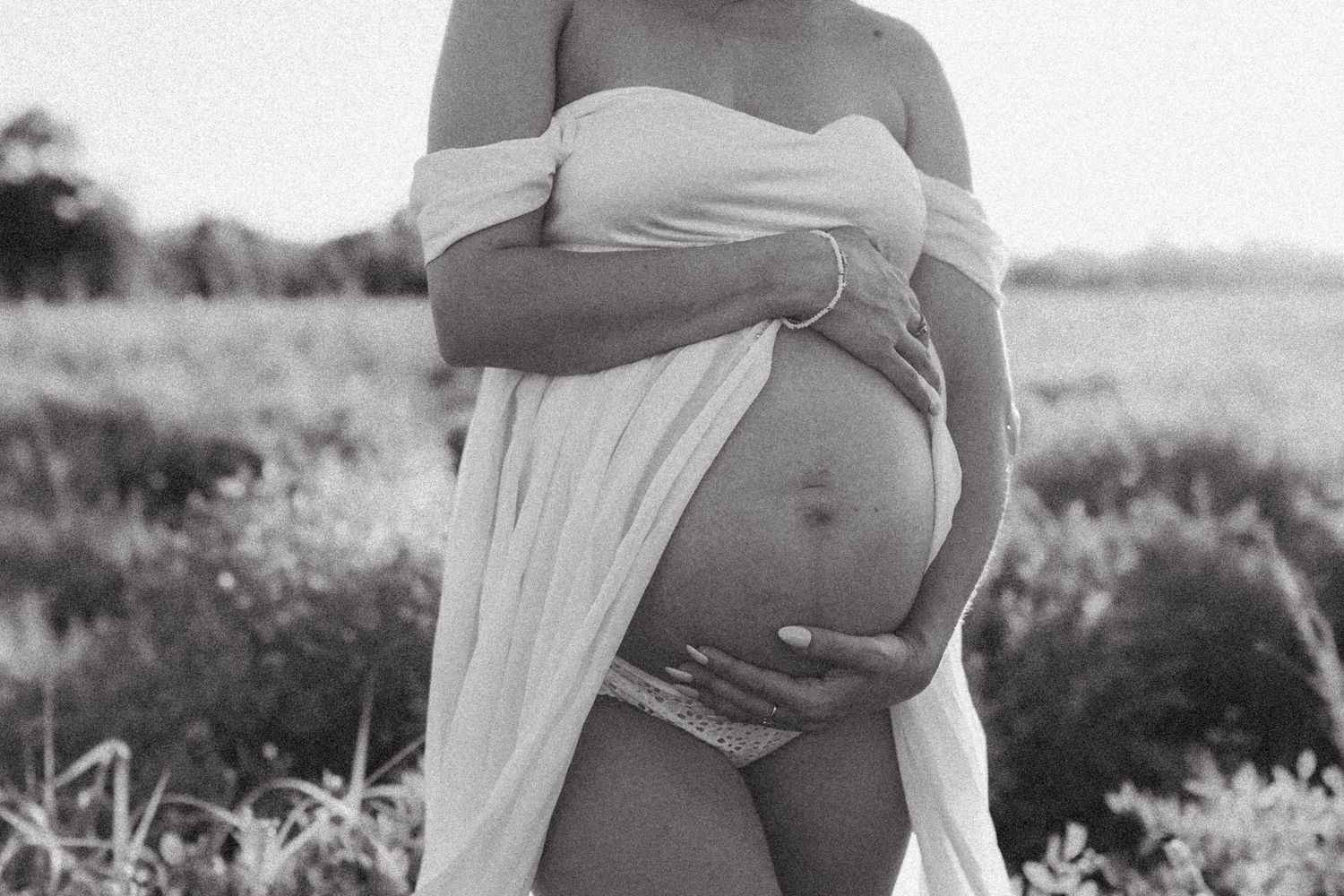 A black and white maternity photo showing a pregnant belly draped in flowing white fabric outdoors in a natural setting.