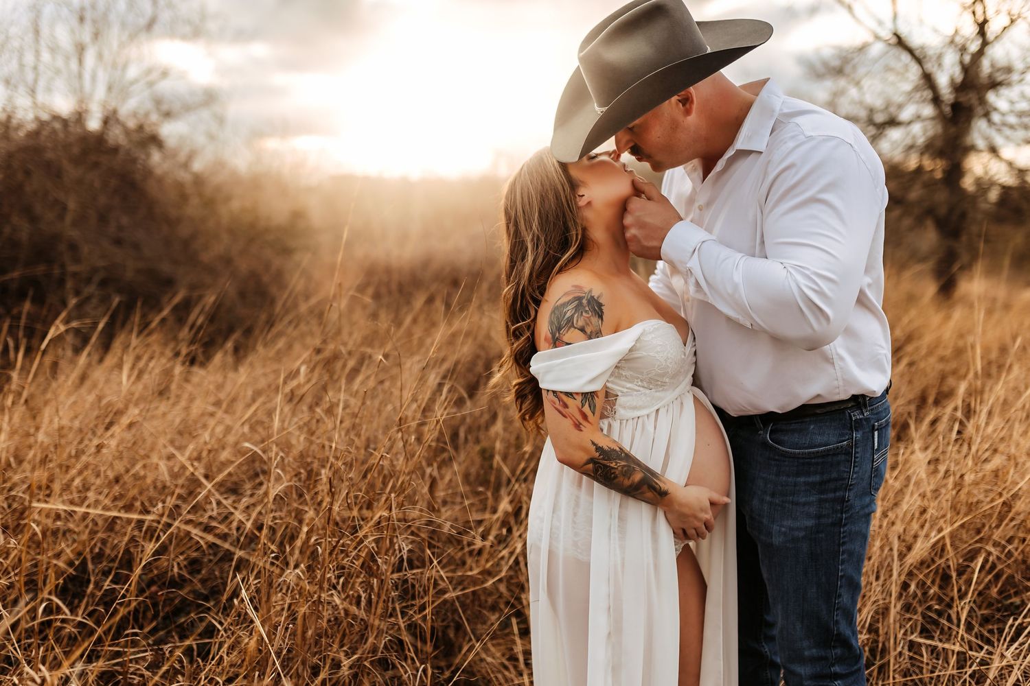 A romantic couple embraces in a golden field at sunset, with the man wearing a cowboy hat and jeans.