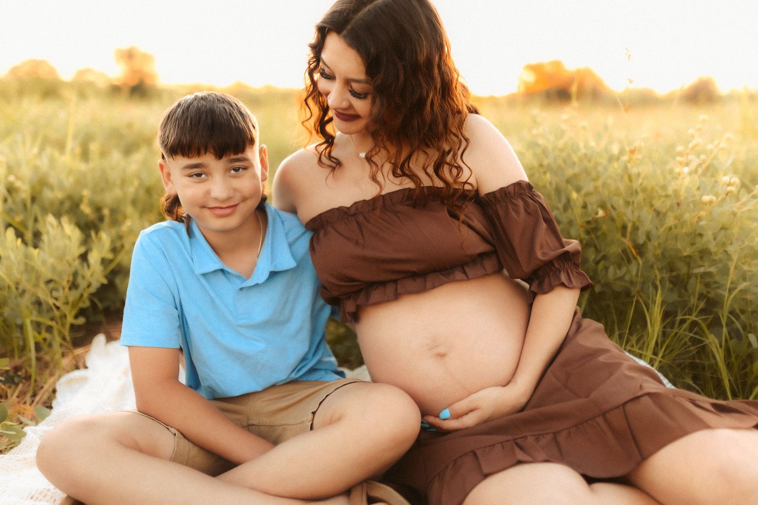 Maternity photo in a golden field at sunset showing mother in brown dress with child sitting in grass.
