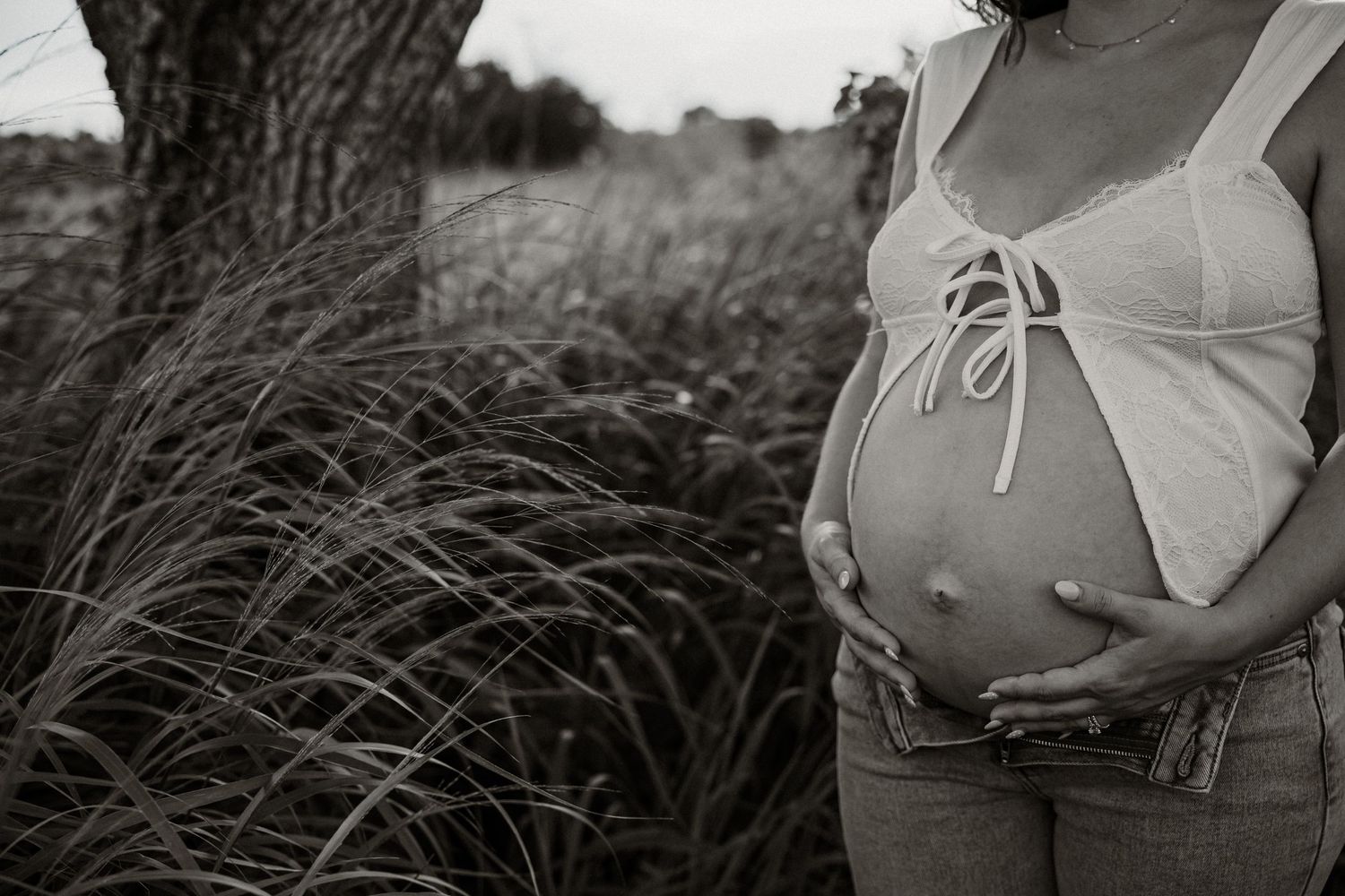 A black and white photograph showing a pregnant belly in a white tank top against a backdrop of tall grass.