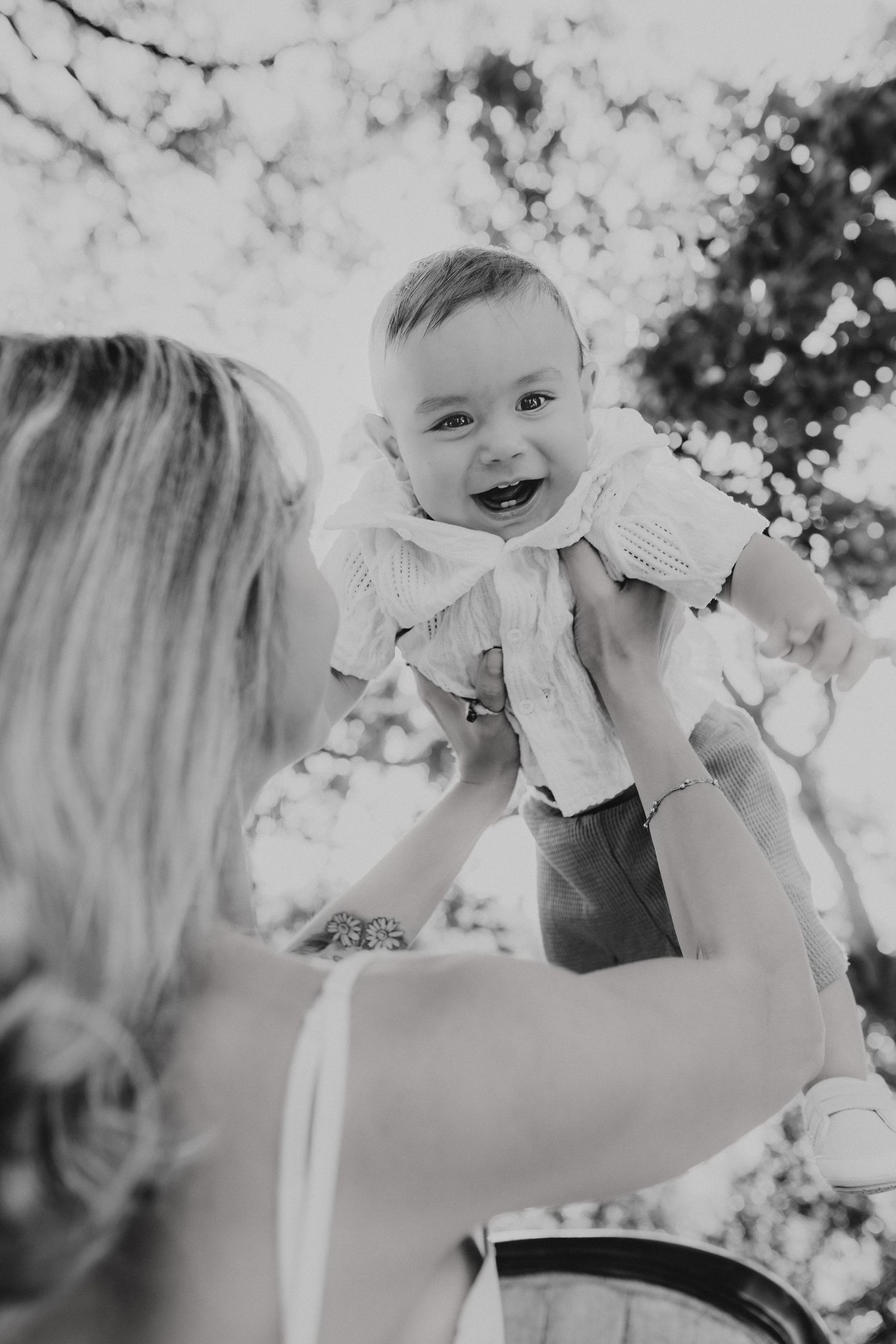 Black and white photo of joyful baby wearing light colored top being lifted in the air outdoors.