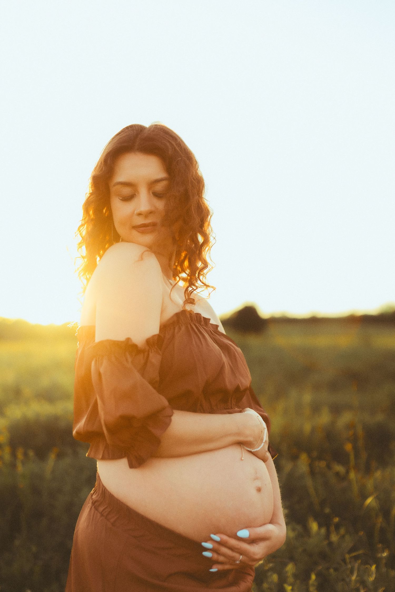 Silhouetted figure holding a pregnant belly against a golden sunset in a natural field setting.