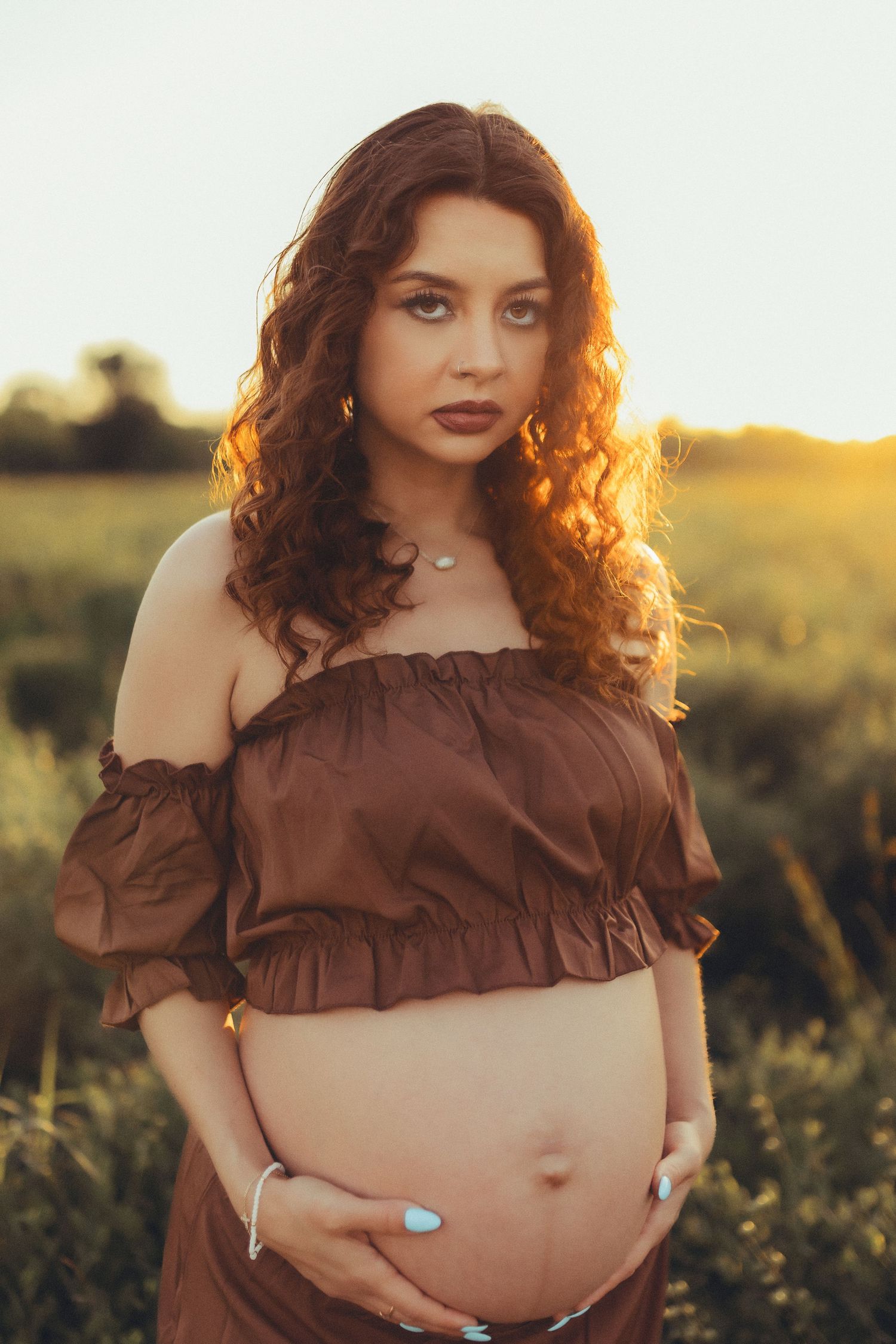 A person in a brown ruffled crop top poses in a field at golden hour with vibrant sunlight in the background.