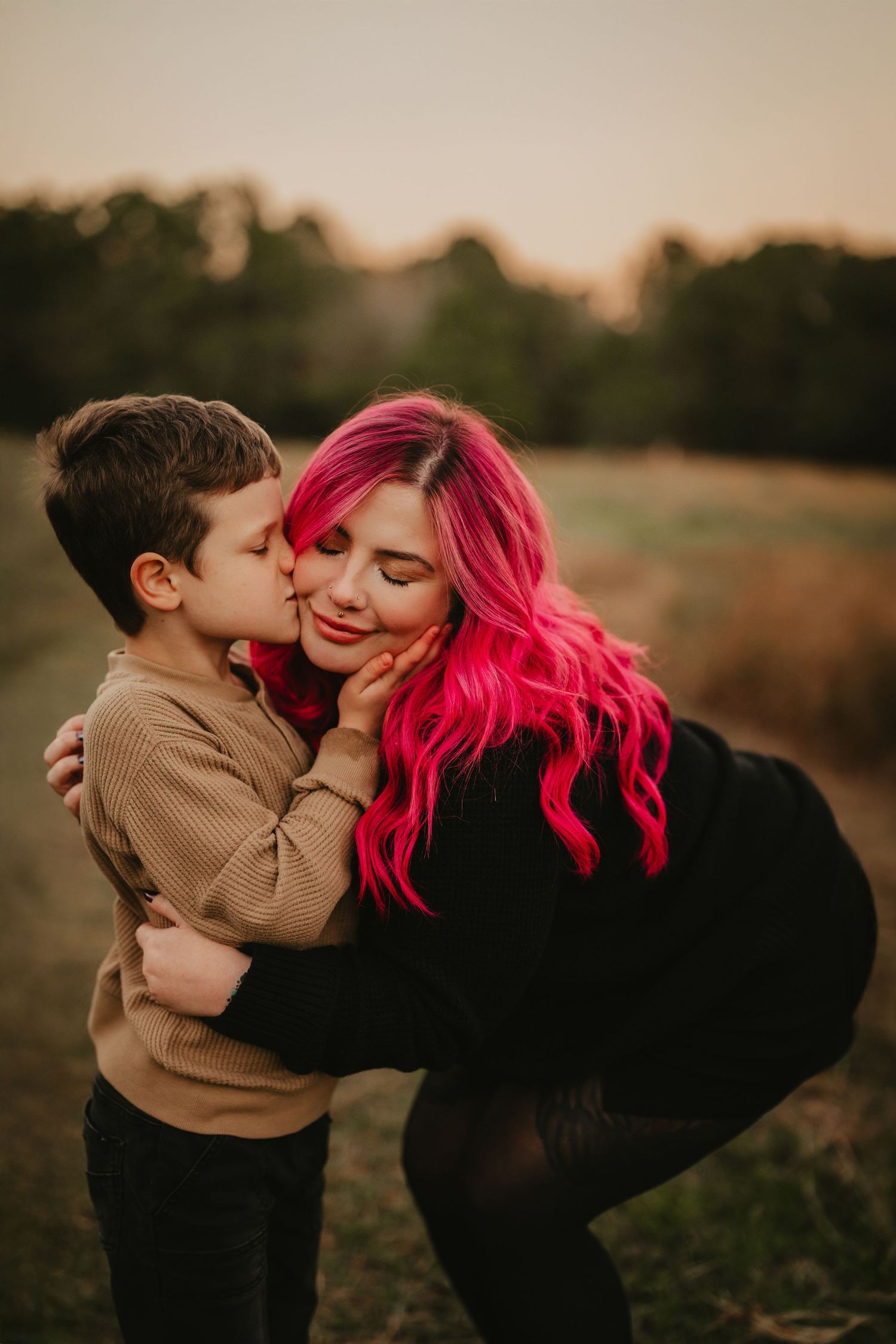 Woman with vibrant pink hair shares a tender outdoor moment with young child in brown sweater at sunset.