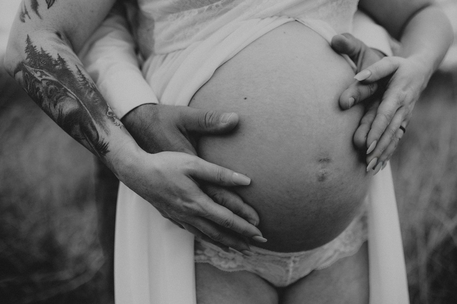 Black and white close-up photograph showing tattooed hands cradling a pregnant belly in soft natural lighting.