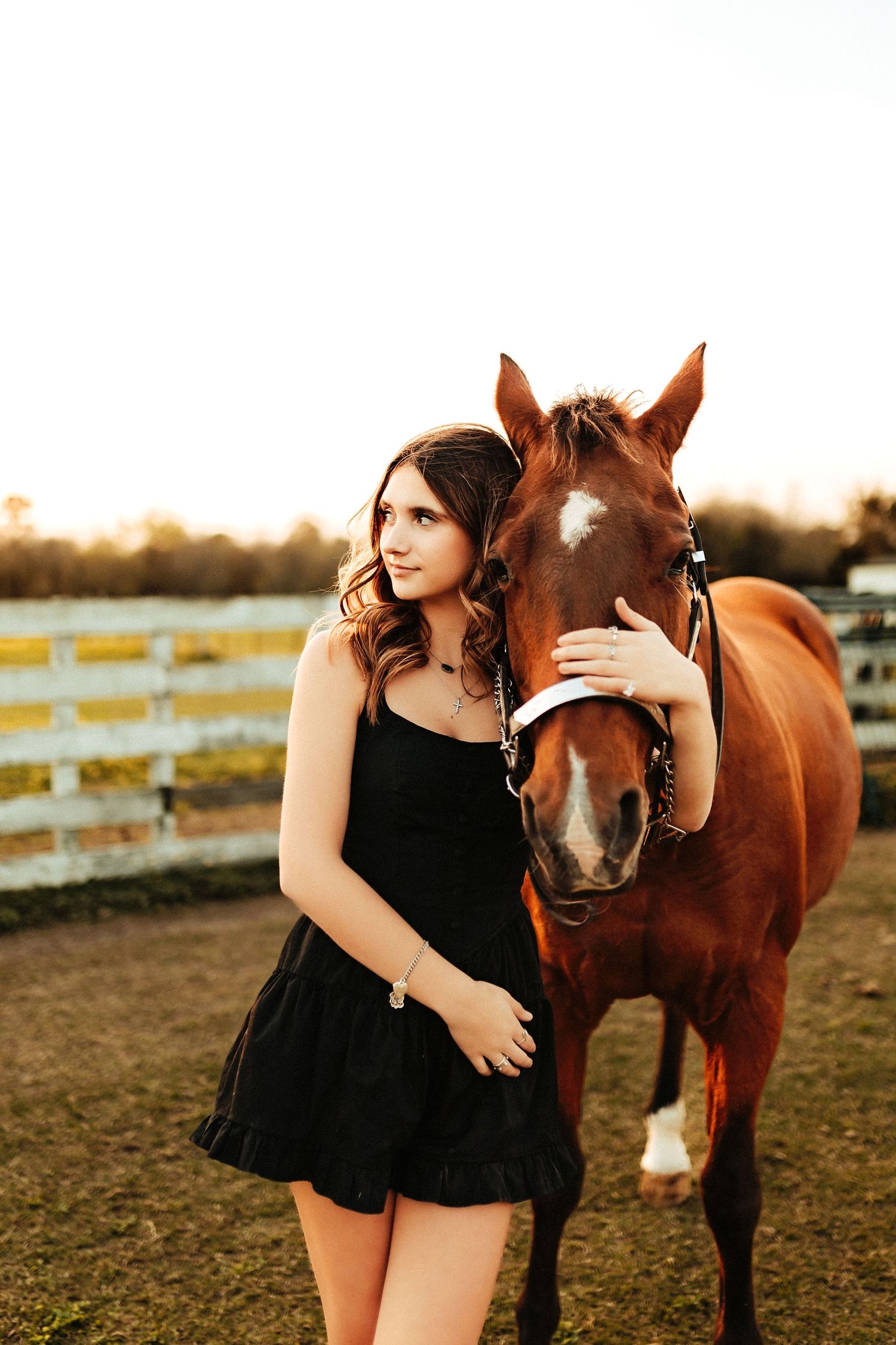 A person in a black dress stands affectionately next to a brown horse in a fenced pasture at sunset.