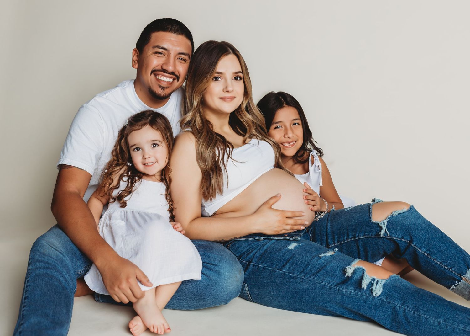 Family members in casual white shirts and jeans pose together on floor against neutral wall in lifestyle photoshoot.