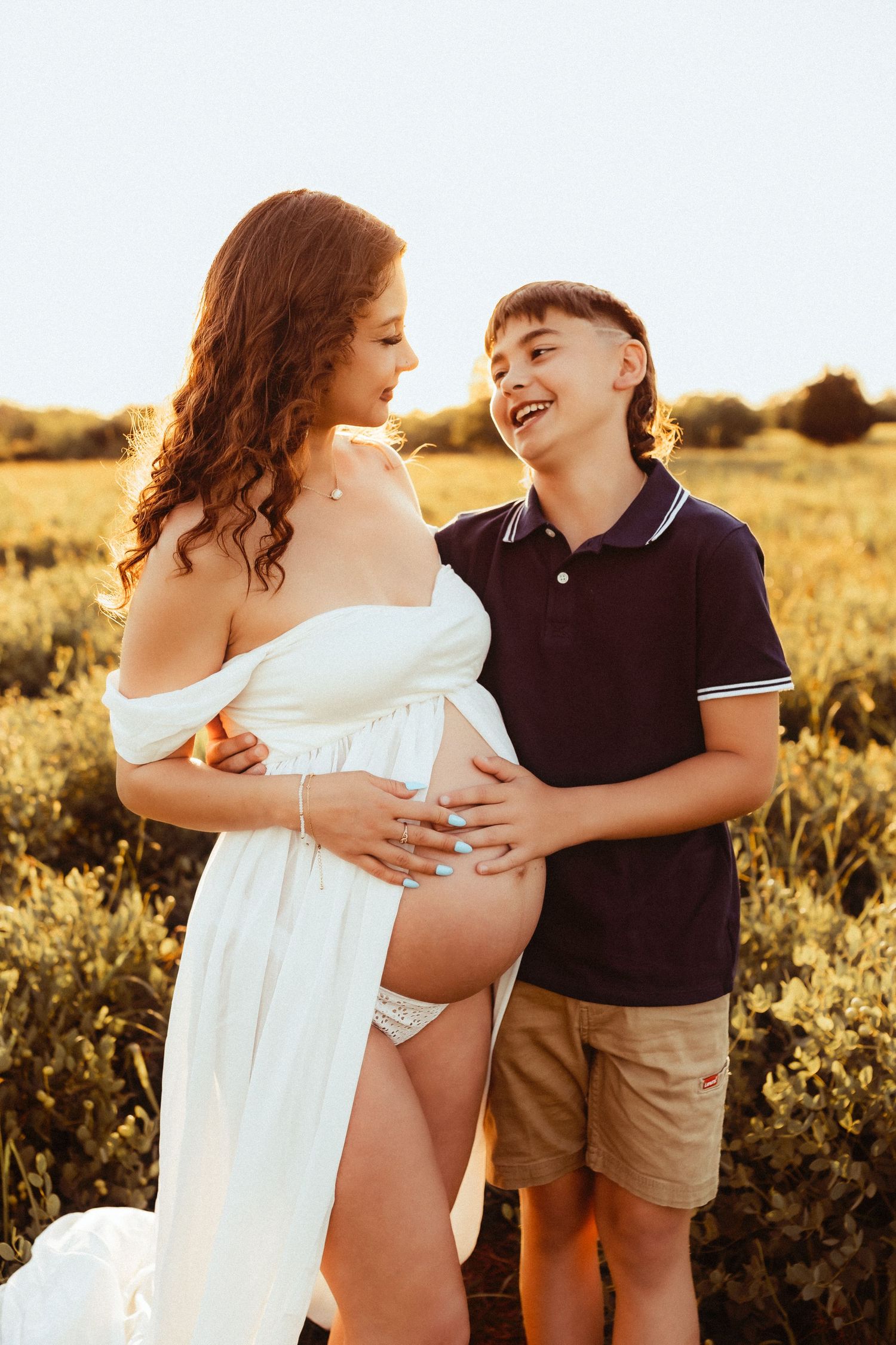 Mother in a flowing white dress embraces her young child in a golden sunlit field during sunset.
