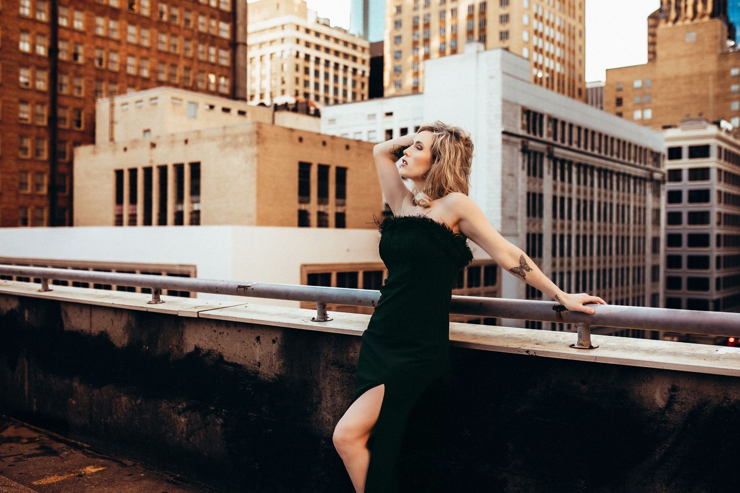 A woman in a black dress poses dramatically on a rooftop with urban architecture in the background.