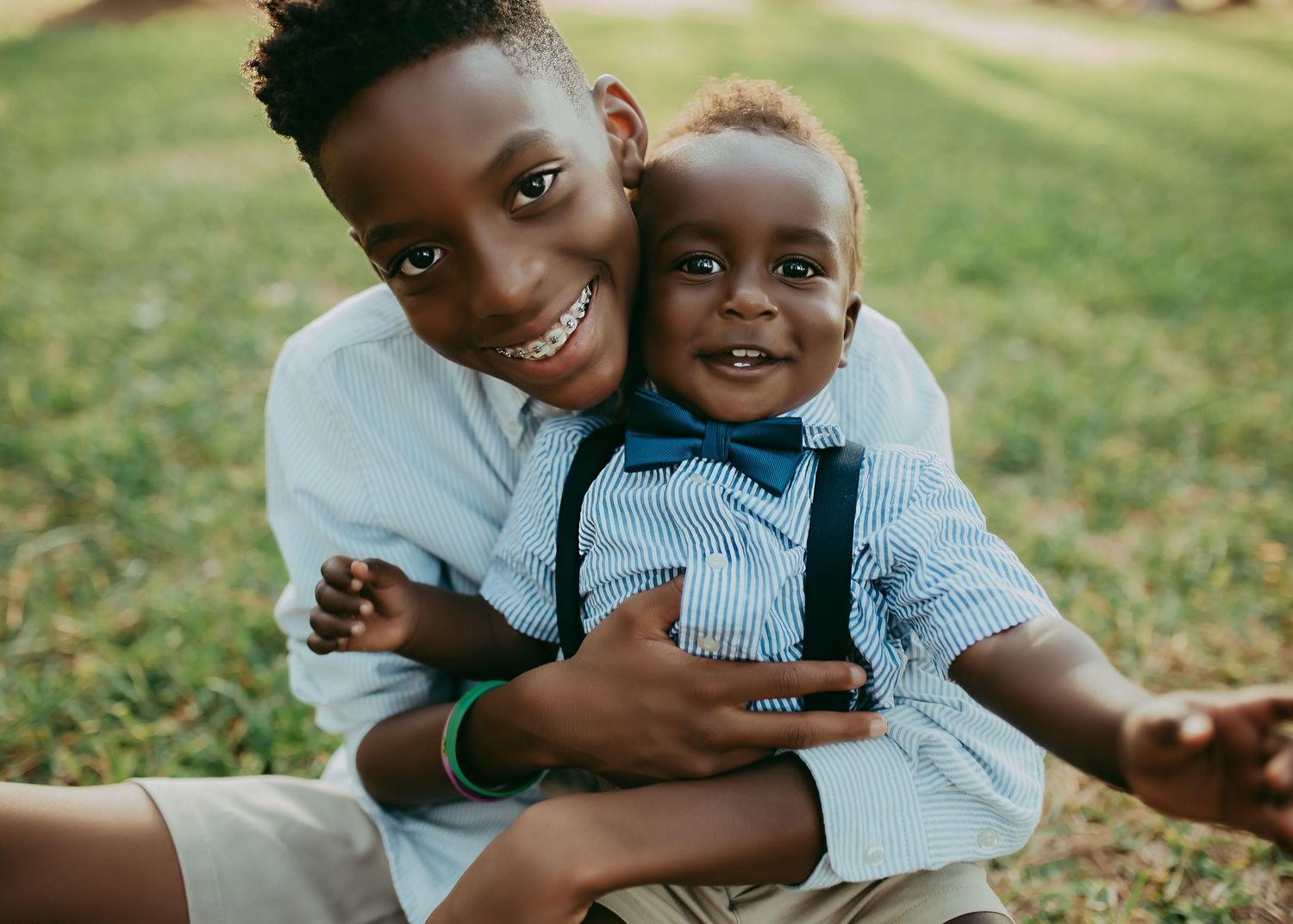 Two people share a joyful embrace while sitting on grass during a sunny outdoor photo session.