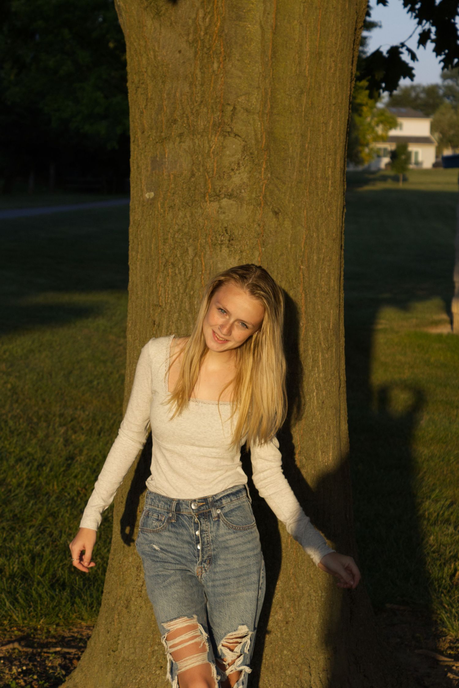 A young woman in a plain white shirt stands against a tree, bathed in the golden hour's lighting.